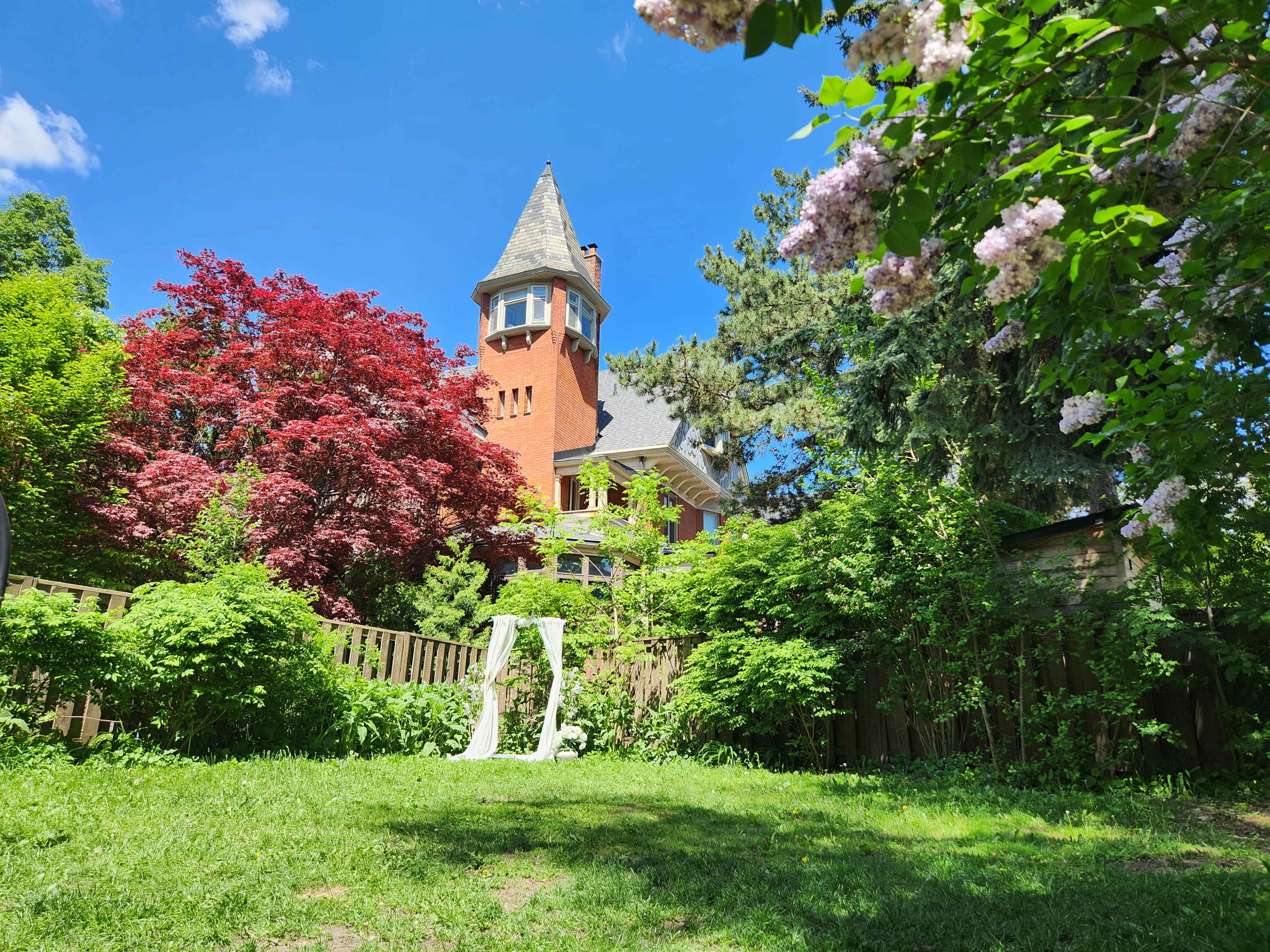 A tall, red brick house with a pointed tower is surrounded by vibrant greenery and colorful trees in a garden setting.