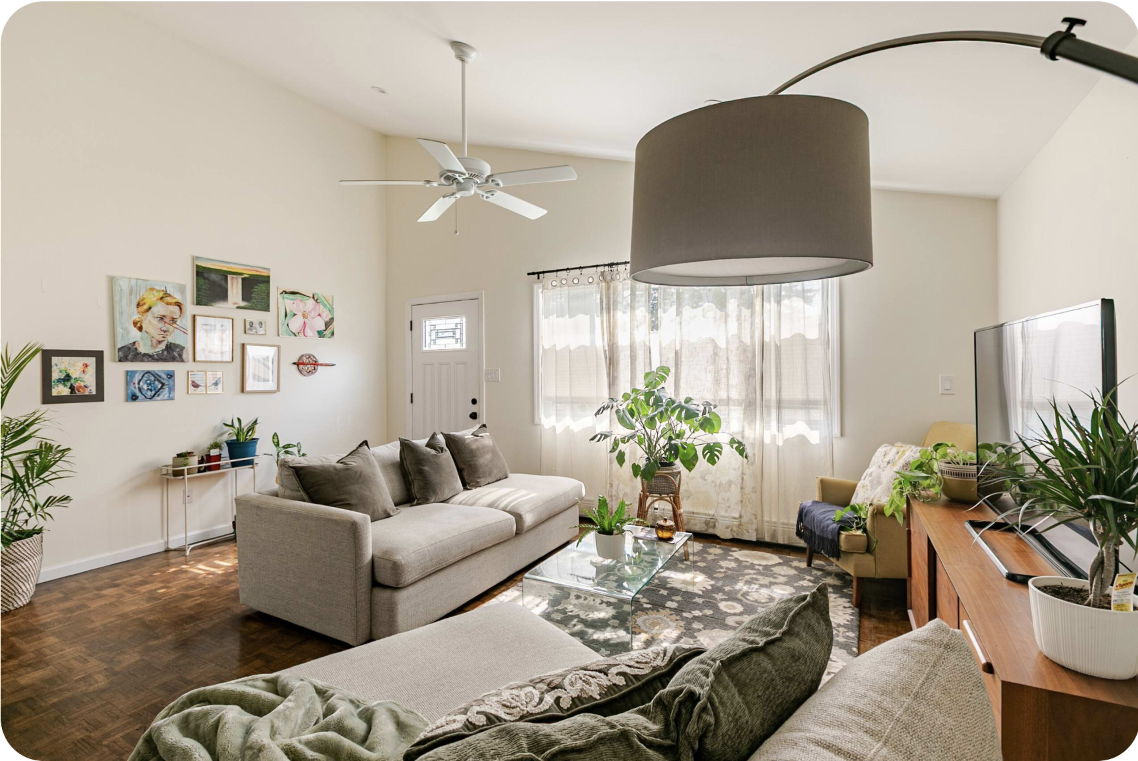 The living room features two sofas, a coffee table, a large window with sheer curtains, and various houseplants.