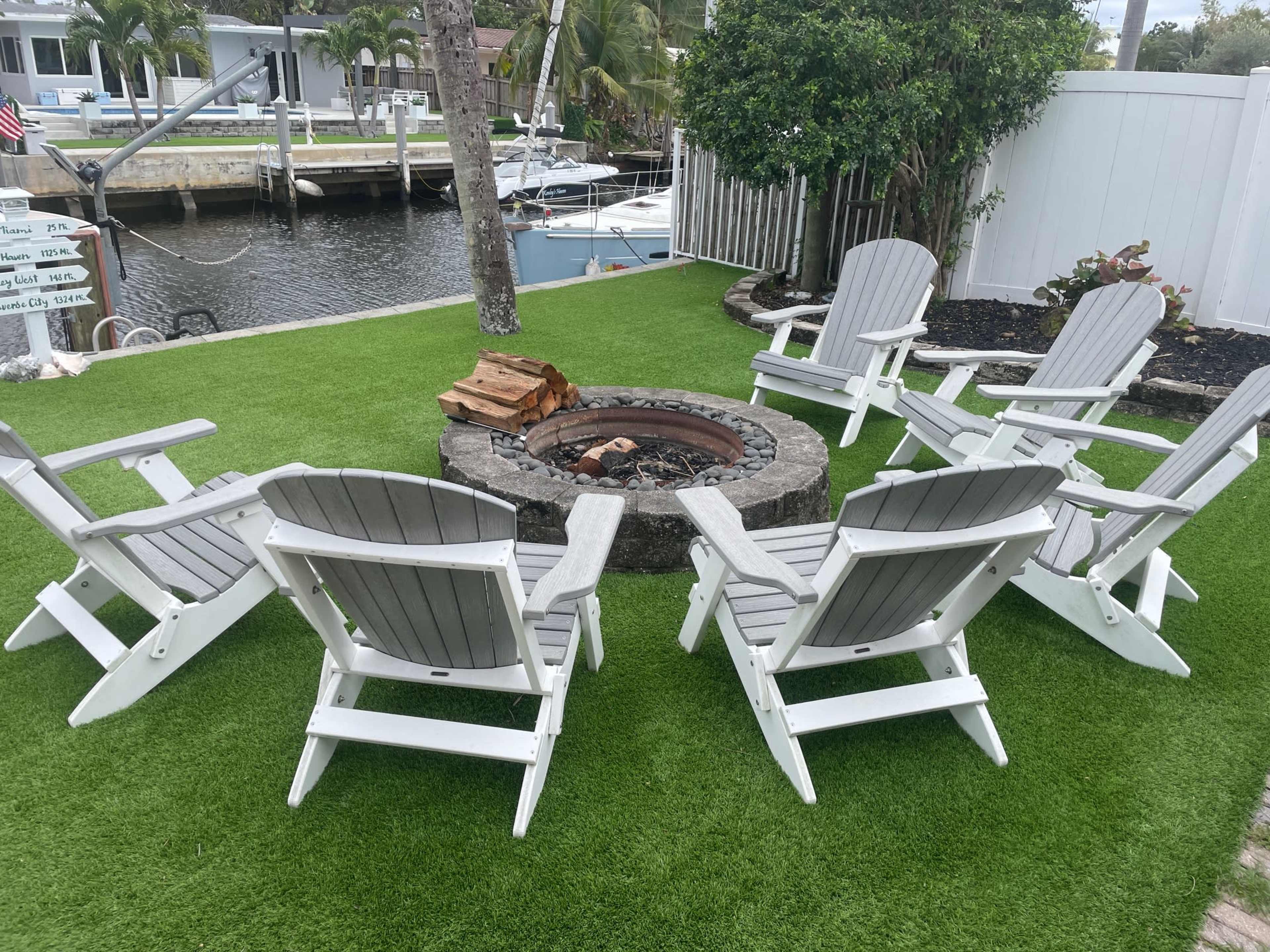 A circular arrangement of white Adirondack chairs surrounds a stone fire pit on a green artificial lawn by a waterway.