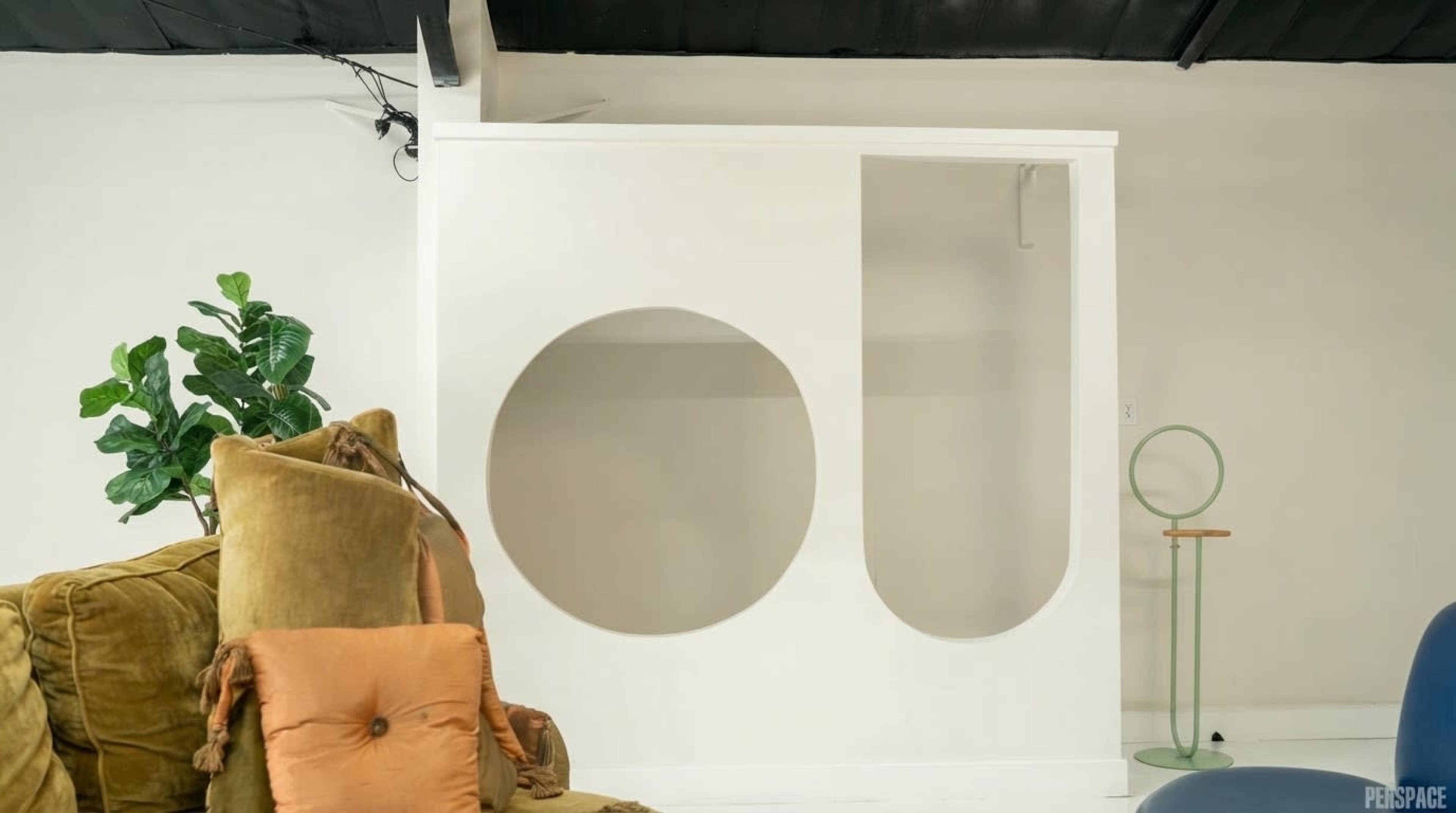 The image shows a minimalistic interior space featuring a white partition with a circular cutout, accompanied by a green plant and a stylish couch with decorative pillows.