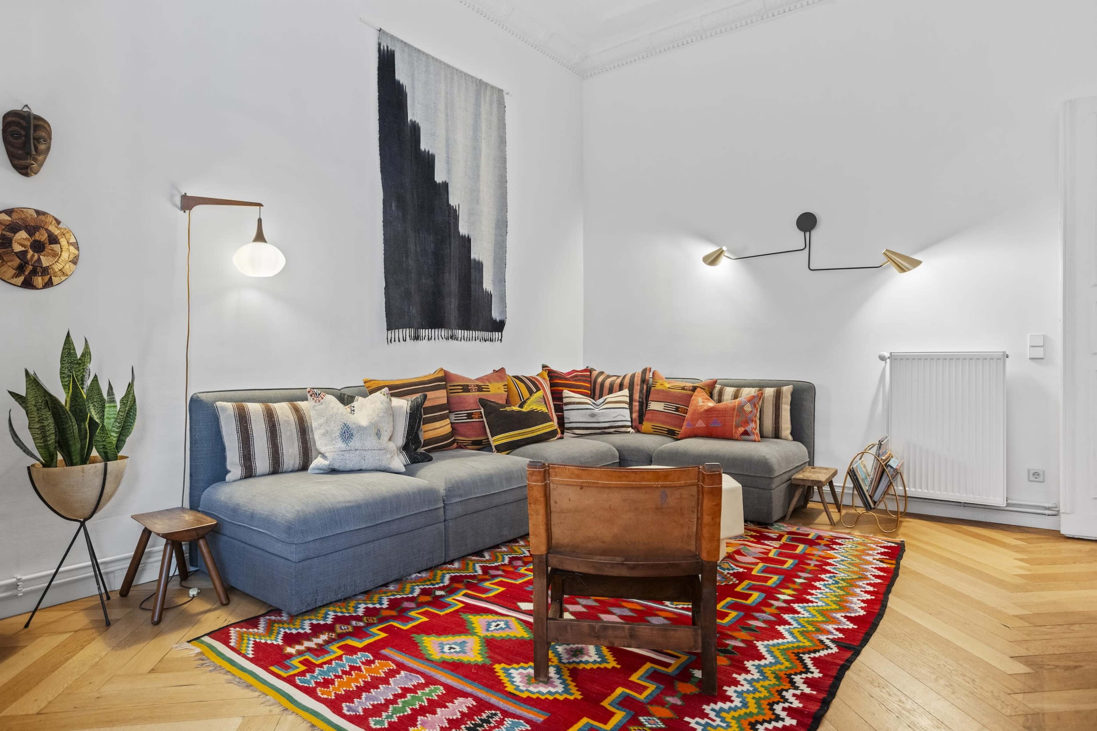 A cozy living room features a sectional sofa adorned with colorful cushions, a patterned rug, and a wall hanging, complemented by plants and decorative wall art.