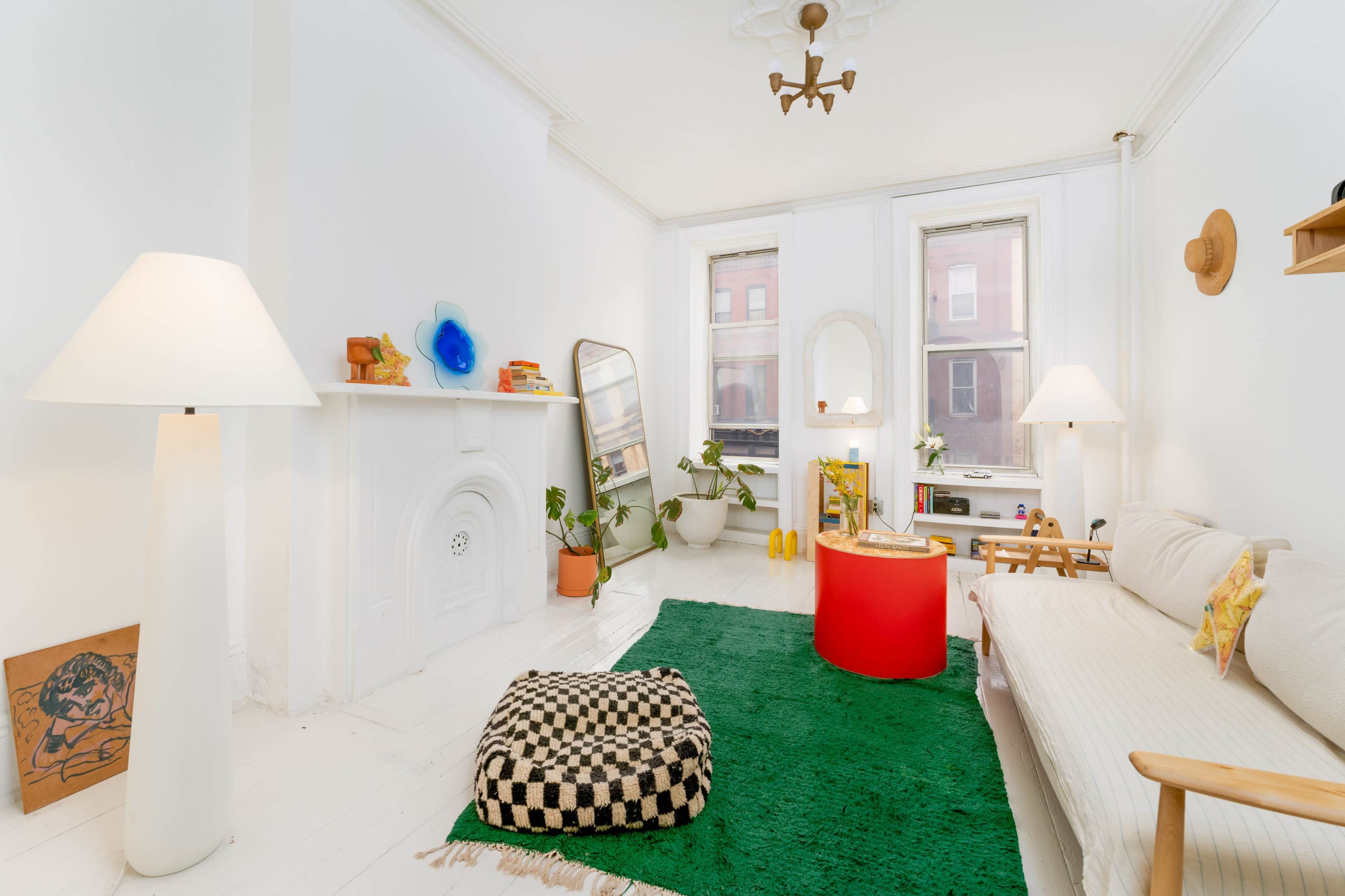 The image shows a bright and minimalist living room with a white sofa, a green rug, a round red table, and large windows allowing natural light.
