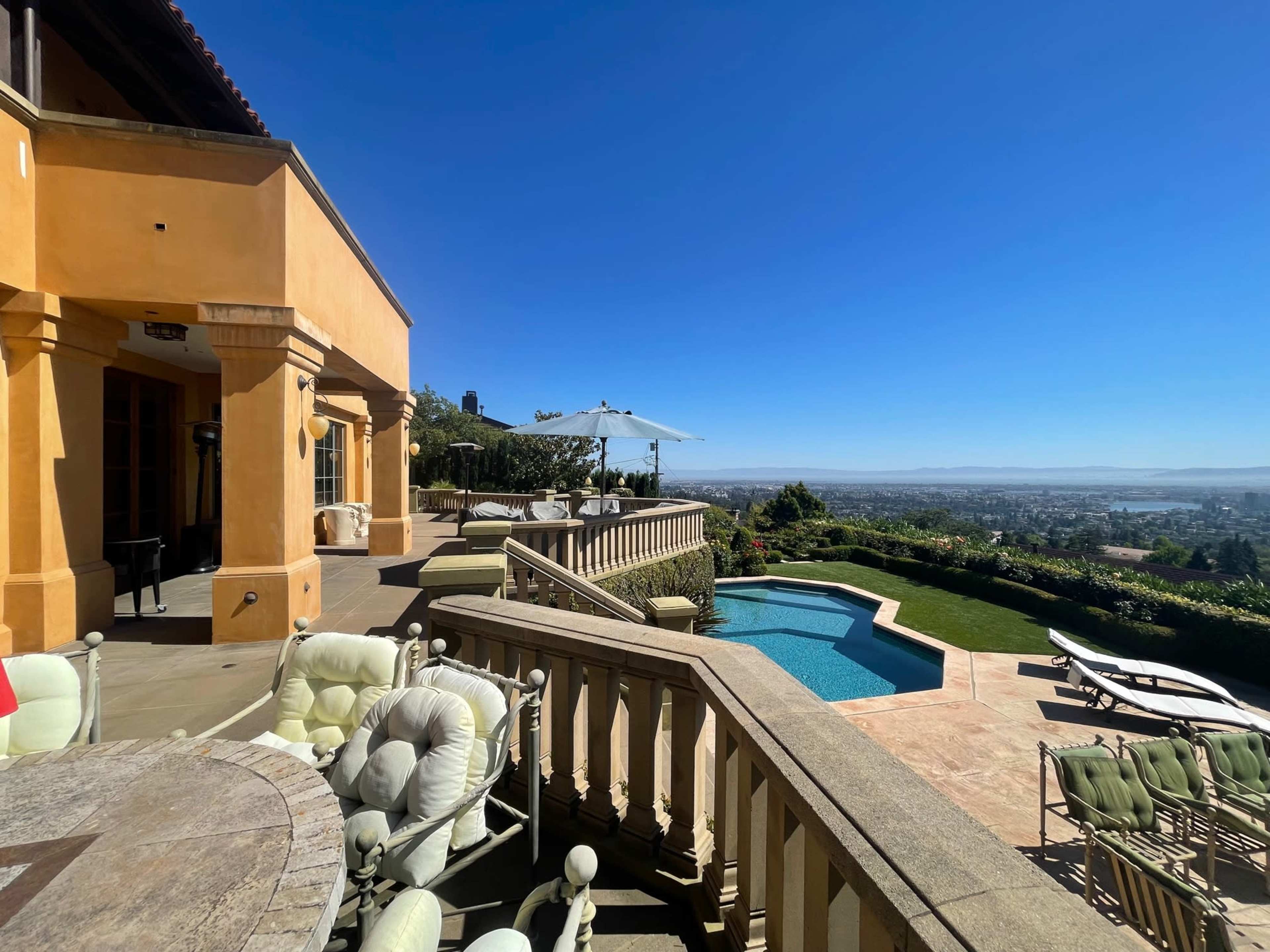 A spacious terrace with outdoor seating surrounds a swimming pool, overlooking a panoramic view of a valley and distant hills under a clear blue sky.