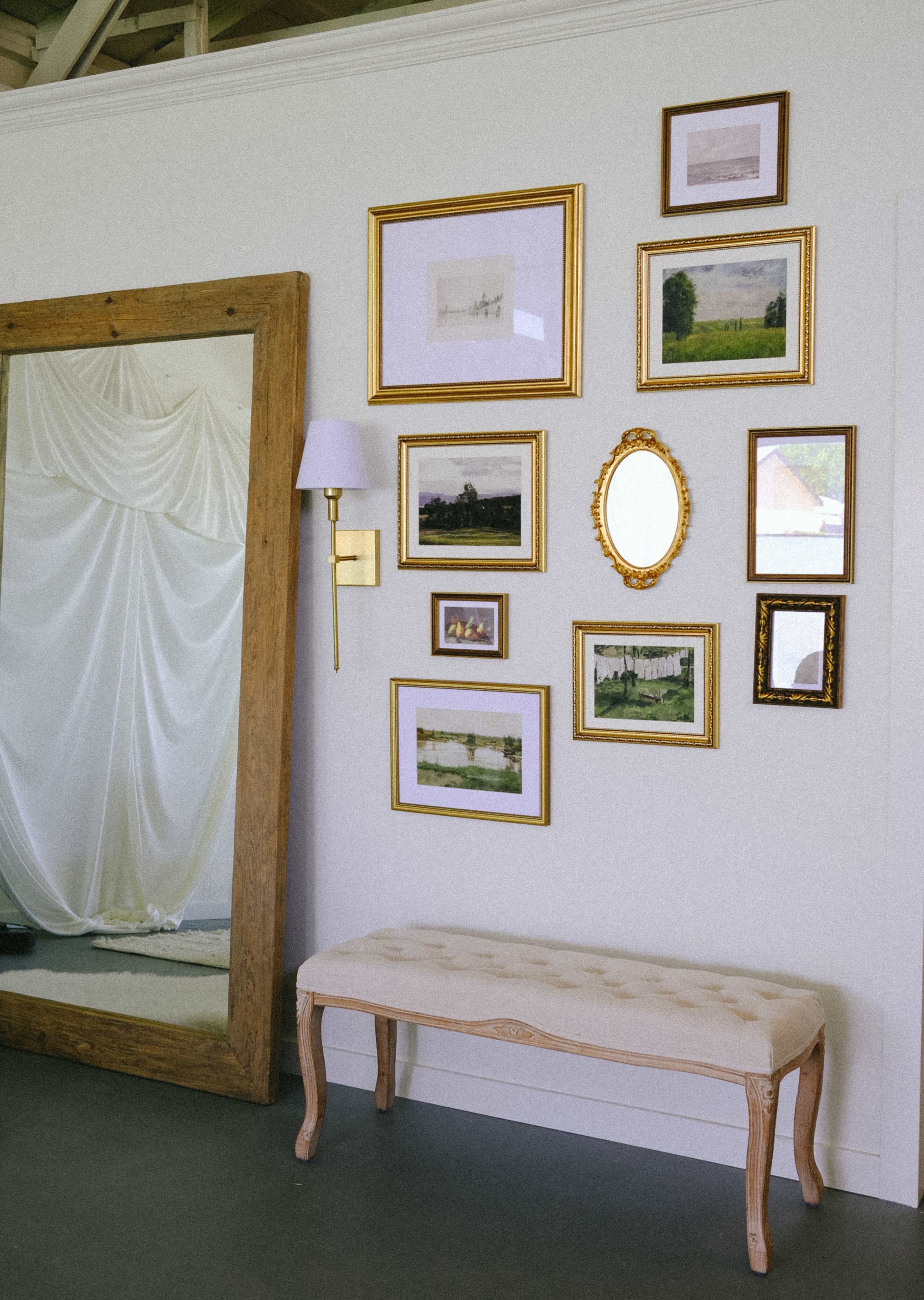 The image shows a wall featuring a collection of framed artwork arranged around a vanity mirror, with a bench in front of it.