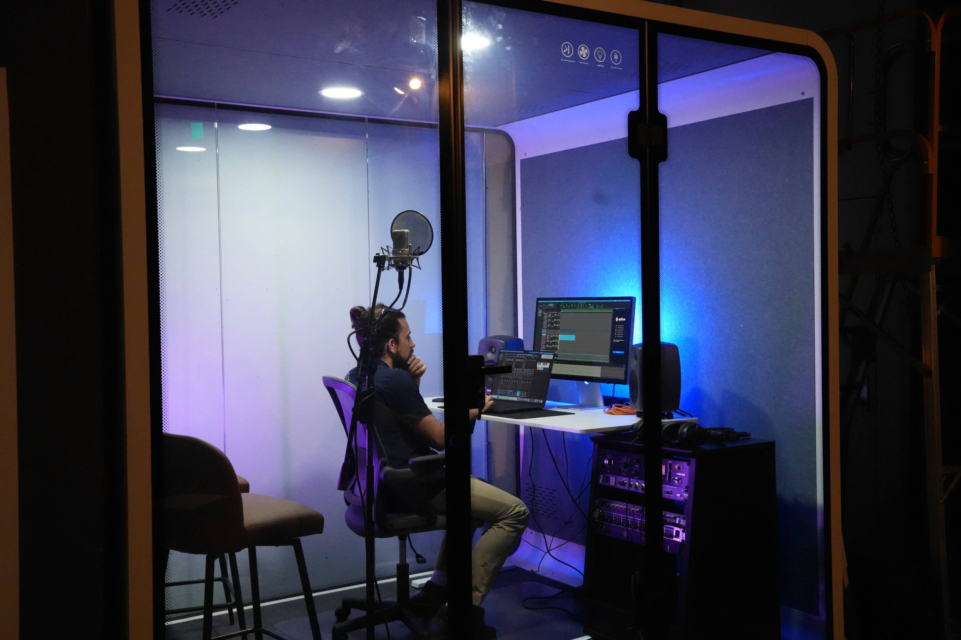 A person sits at a computer in a soundproof booth, with a microphone and various audio equipment visible.