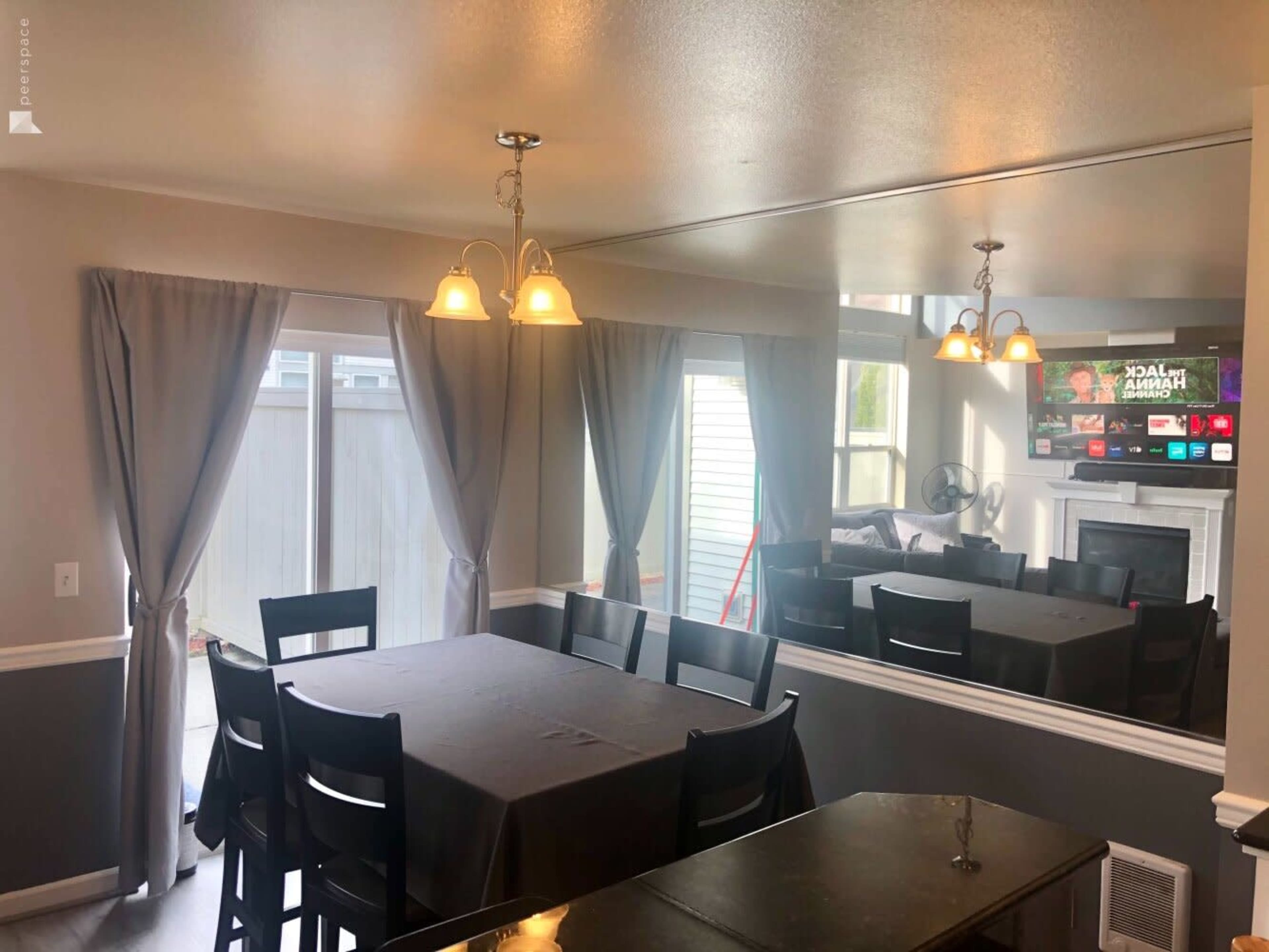 A dining area features a table surrounded by black chairs, with curtains framing a window and a mirrored wall reflecting light and furniture.