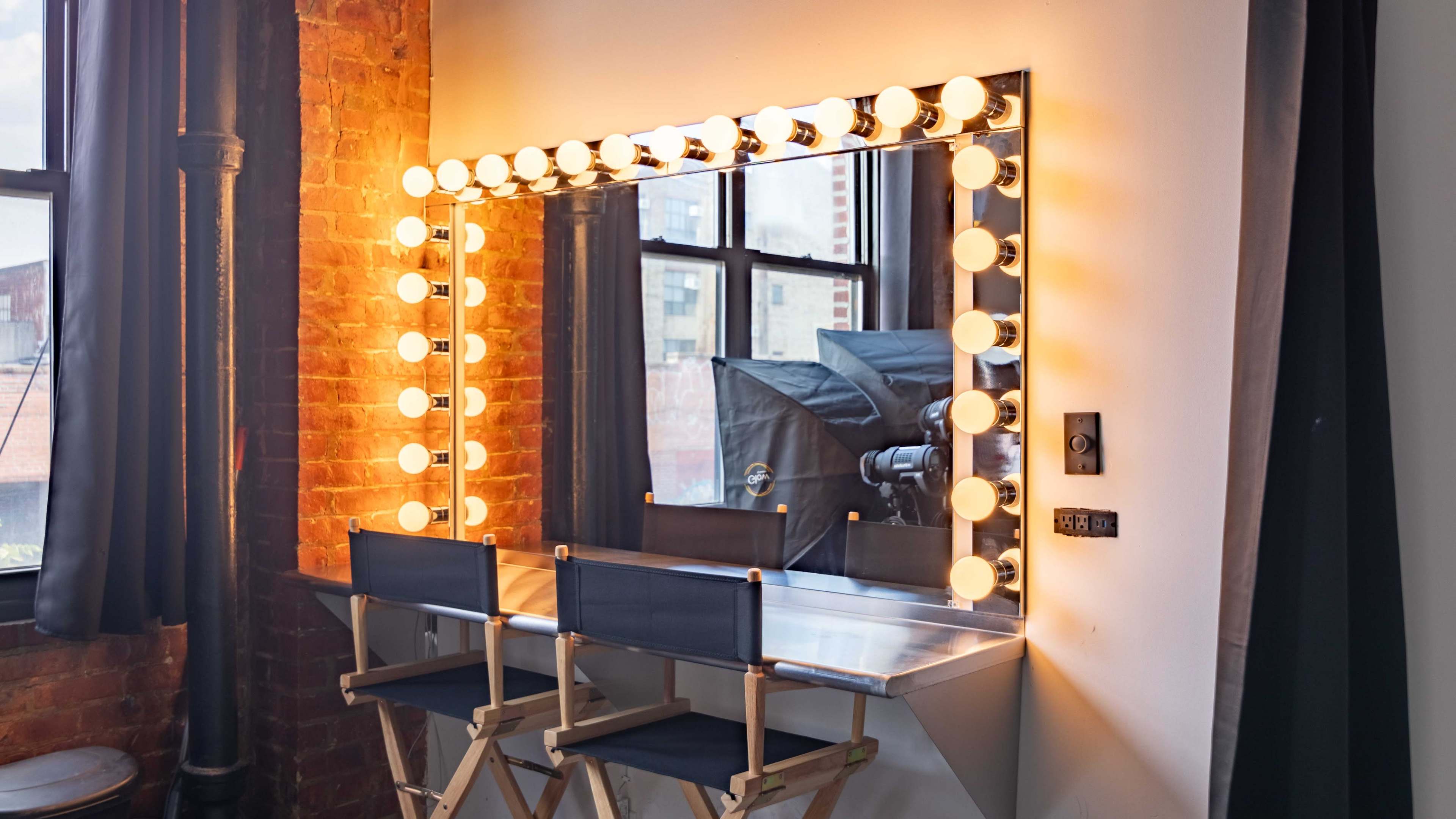 The image shows a well-lit dressing table with a large mirror surrounded by light bulbs, flanked by two director's chairs, set against a brick wall and large windows.