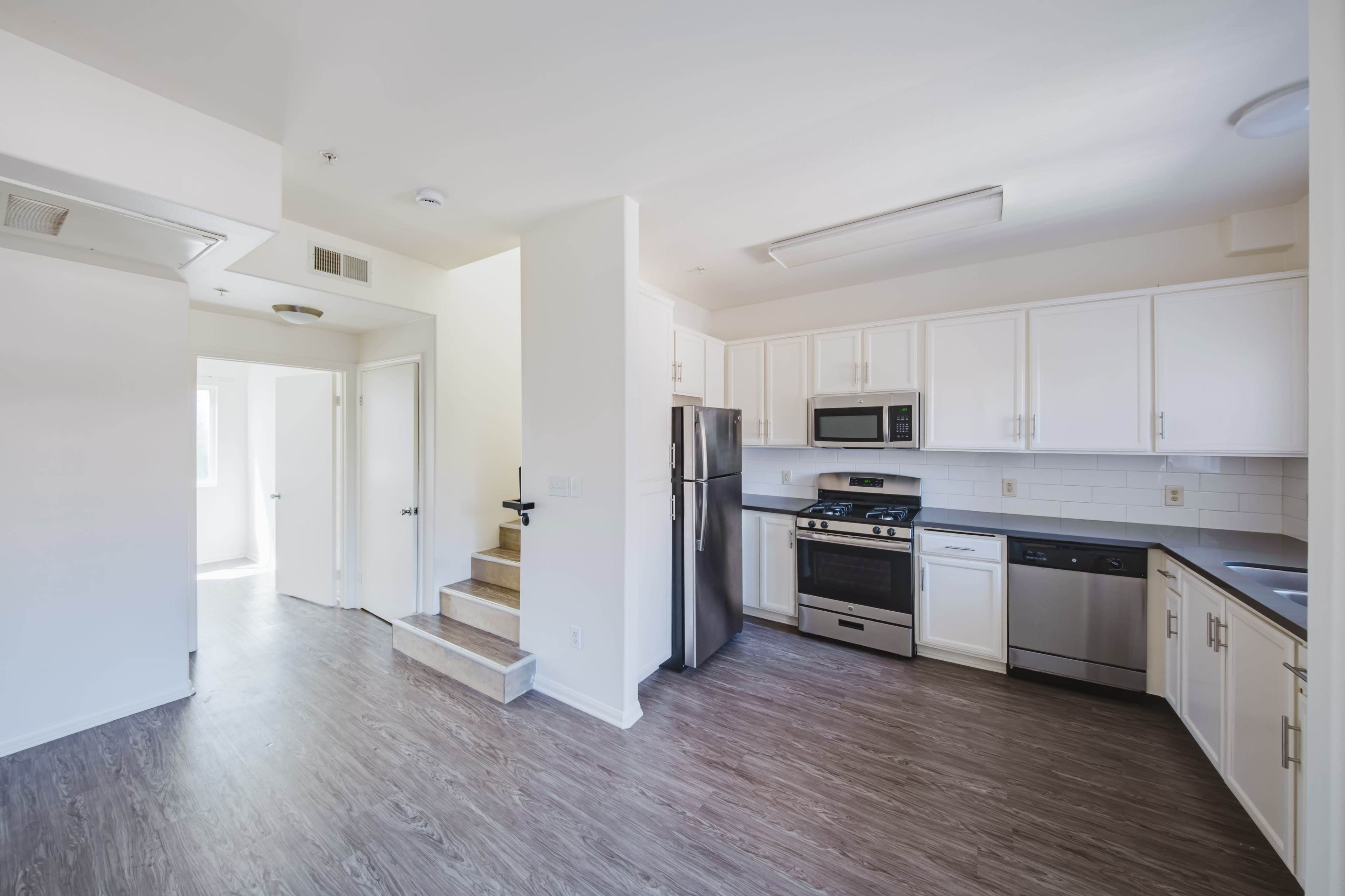 This image depicts a modern kitchen with stainless steel appliances, white cabinetry, and a staircase leading to another room.