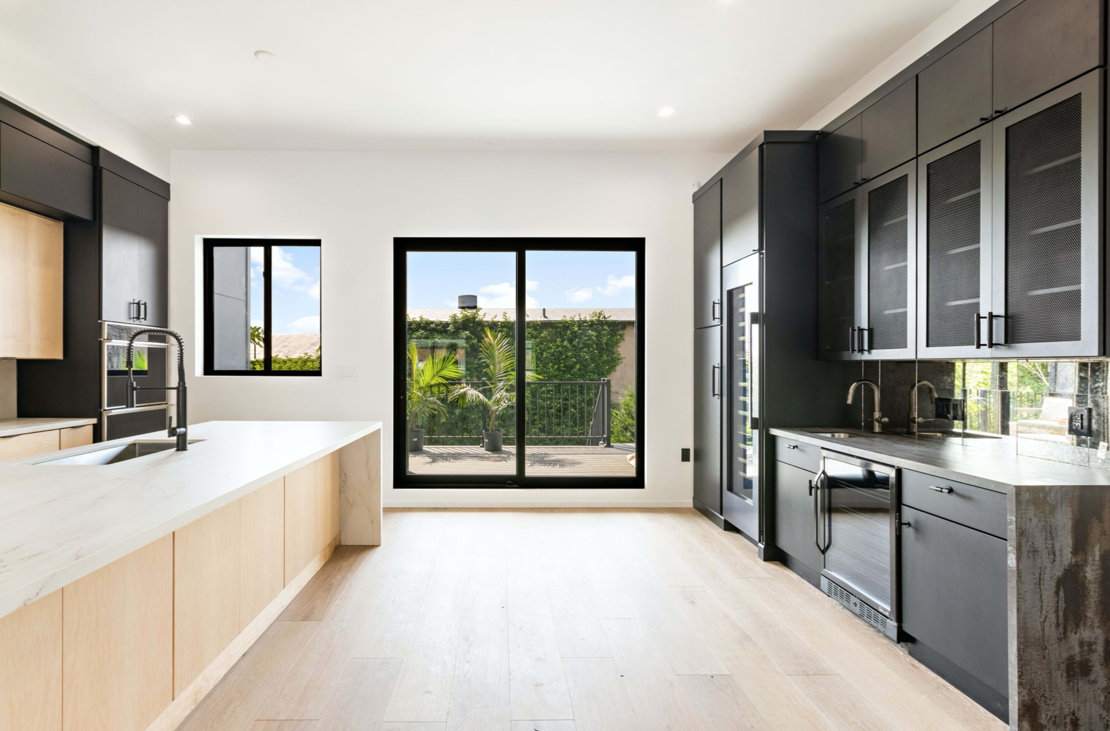 A modern kitchen with black cabinets, a large island, and a view of an outdoor patio through a large window.