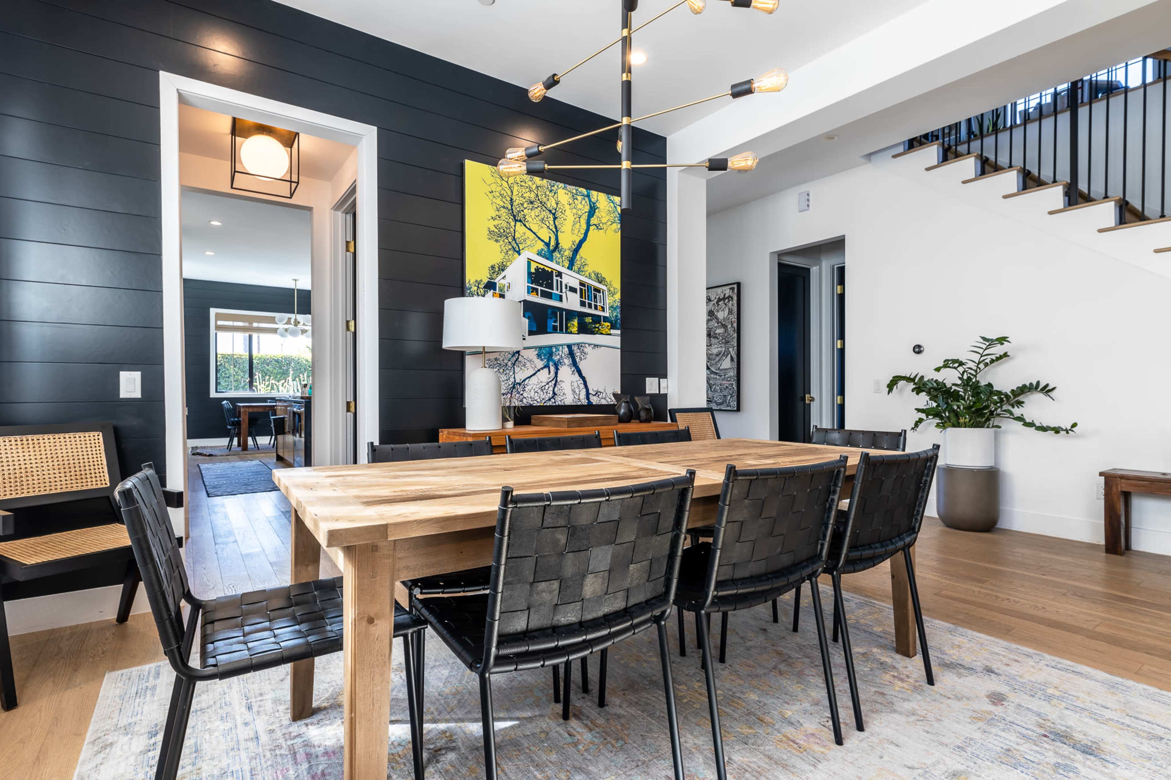 A dining area with a wooden table surrounded by black chairs, featuring a modern chandelier and a colorful artwork on the wall.