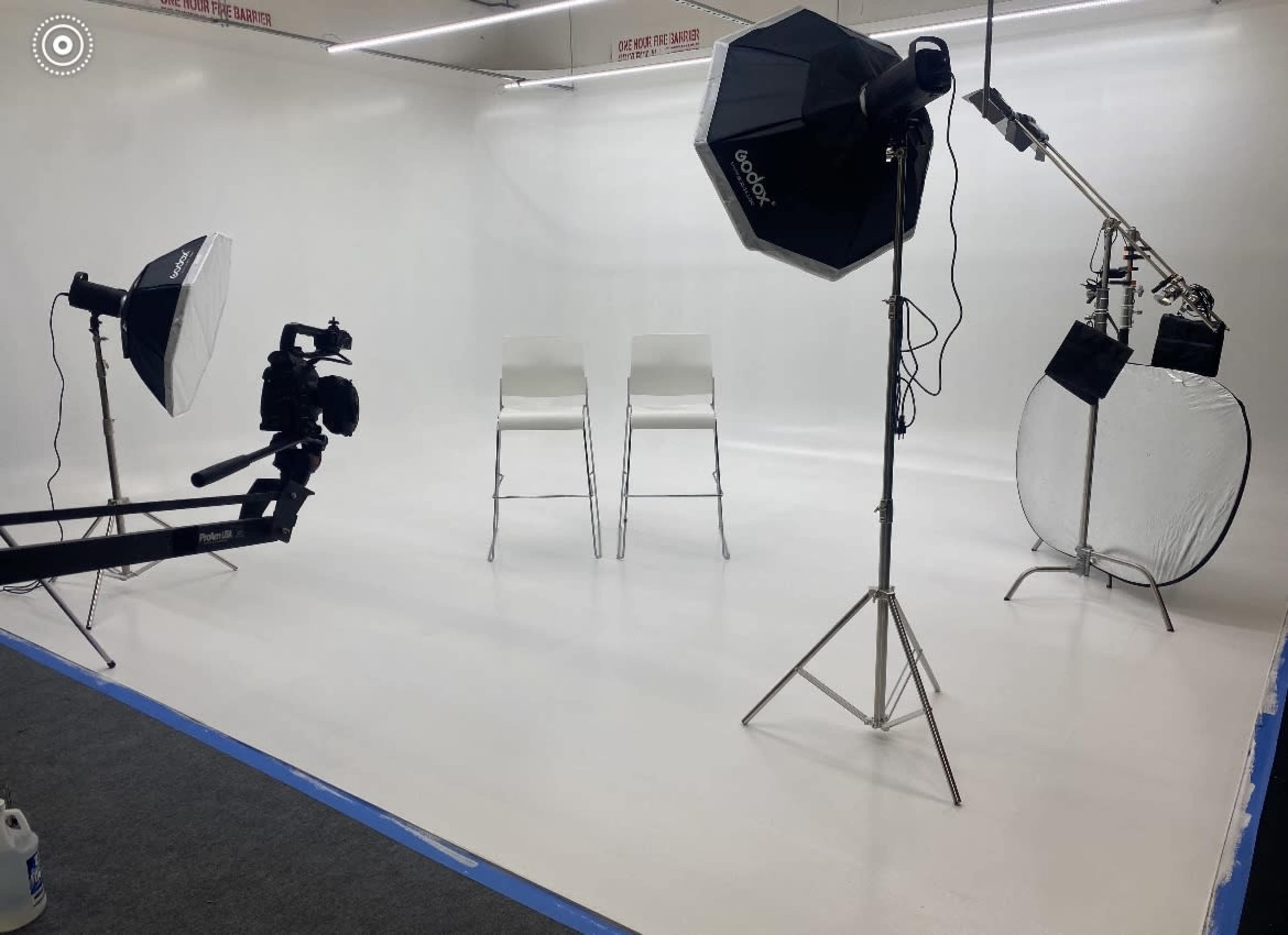 A photography studio with two empty chairs, lighting equipment, and a camera setup against a plain white backdrop.