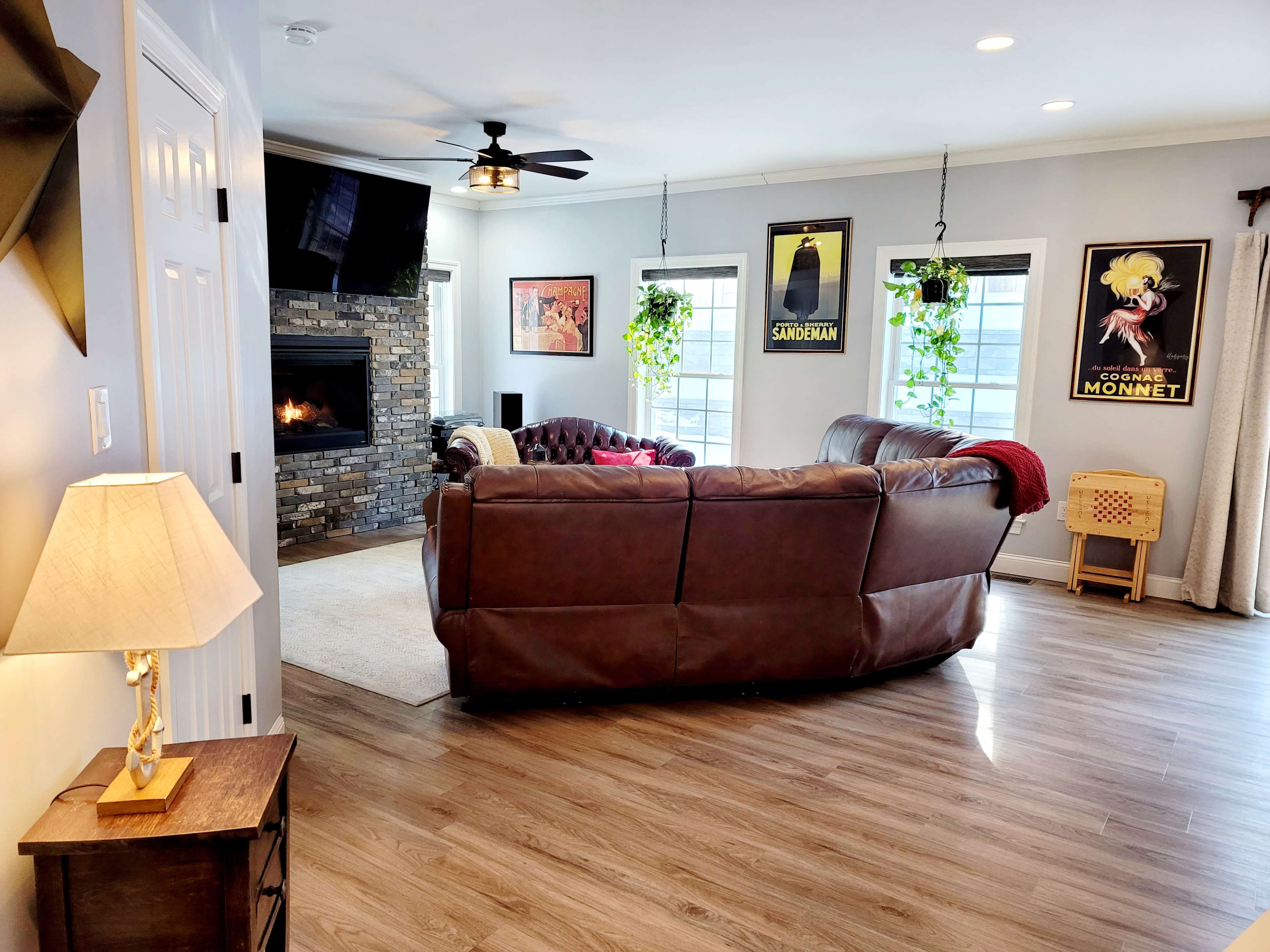 A cozy living room features a brown leather sofa, a stone fireplace, and framed artwork on the walls, with plants hanging from the ceiling.