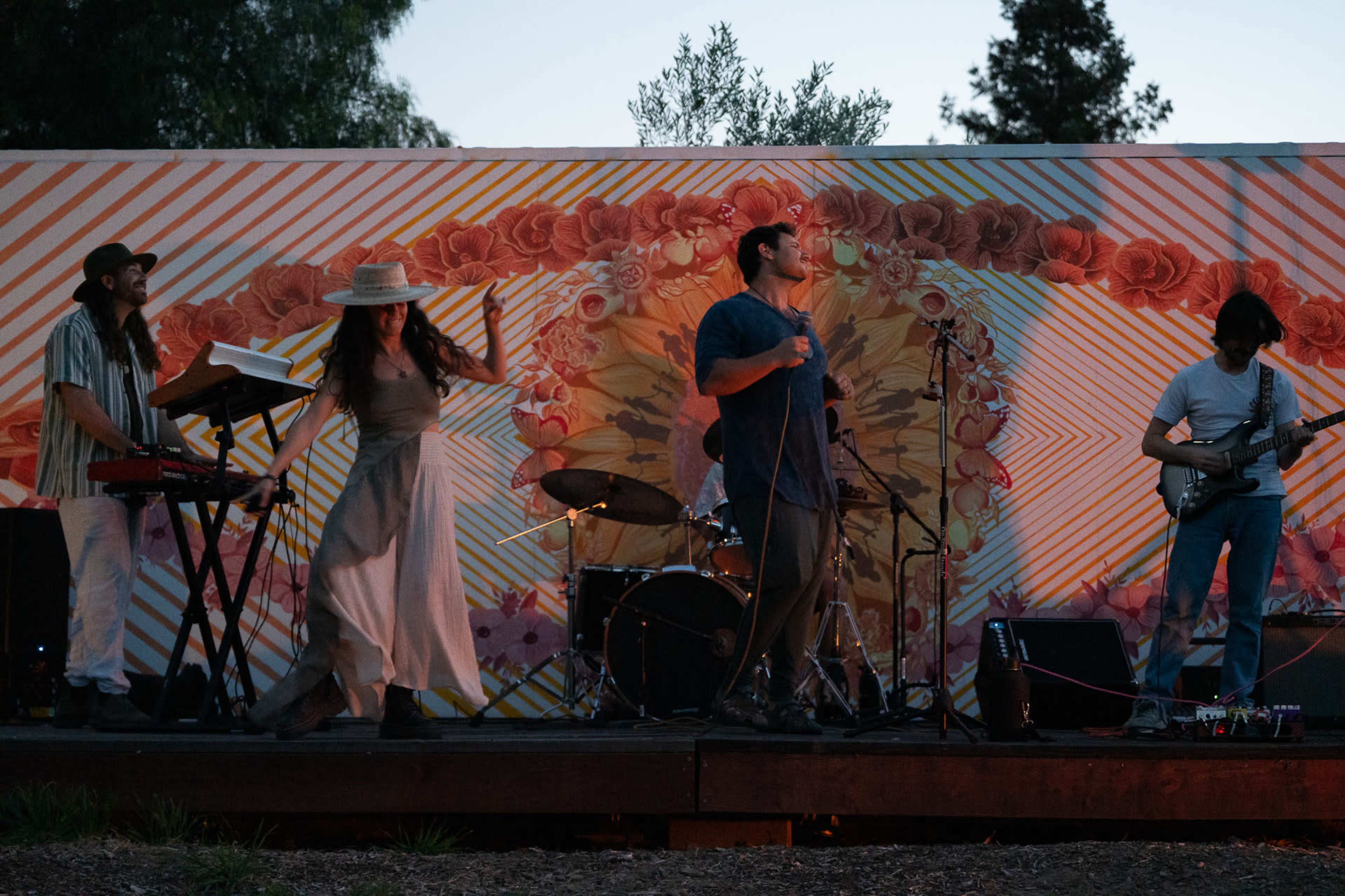 A band performs on stage in front of a colorful mural as the sun sets.