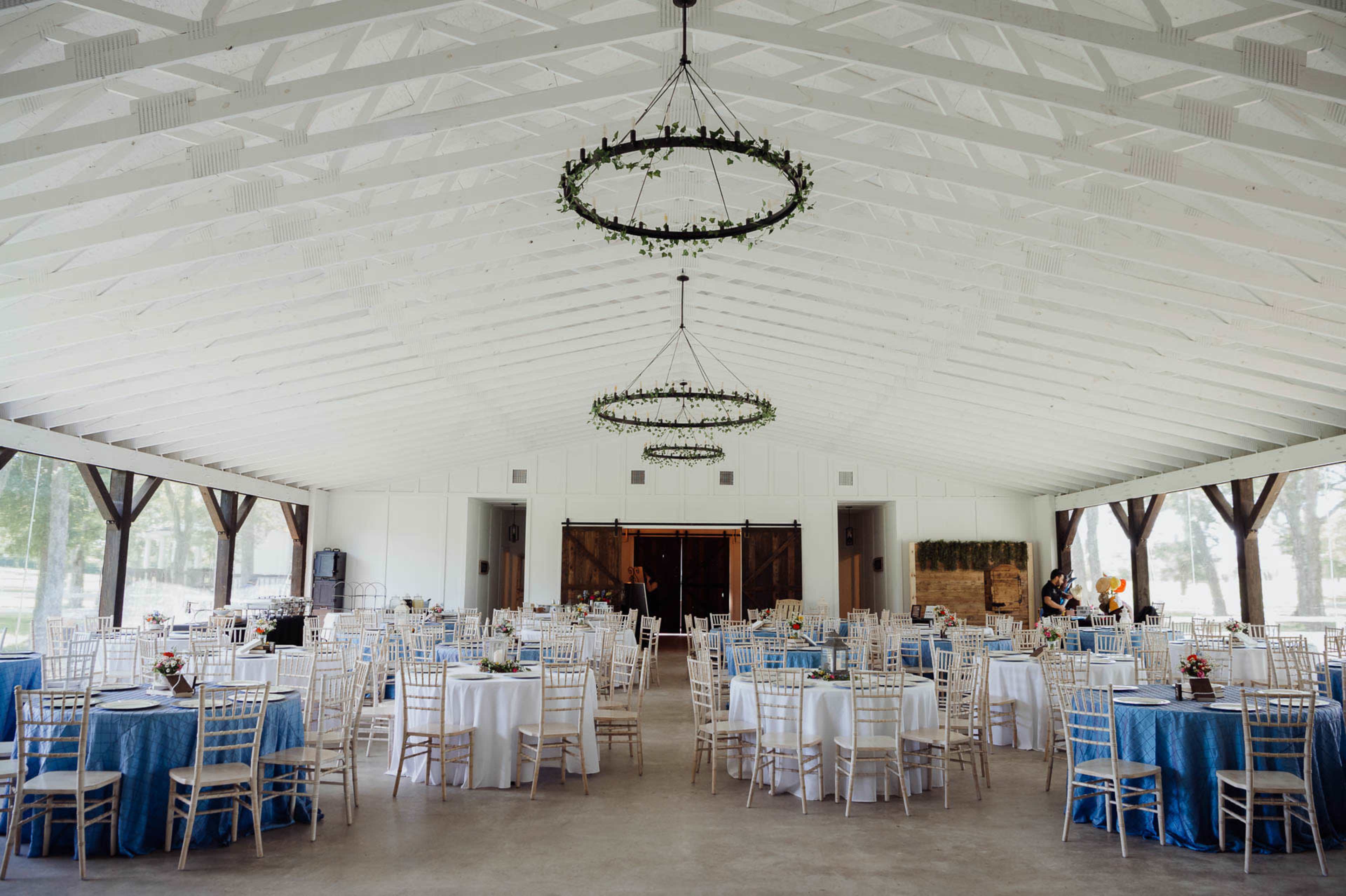 The interior of a spacious, well-lit event hall features round tables with blue tablecloths and white chairs arranged neatly, along with two circular chandeliers hanging from the ceiling.