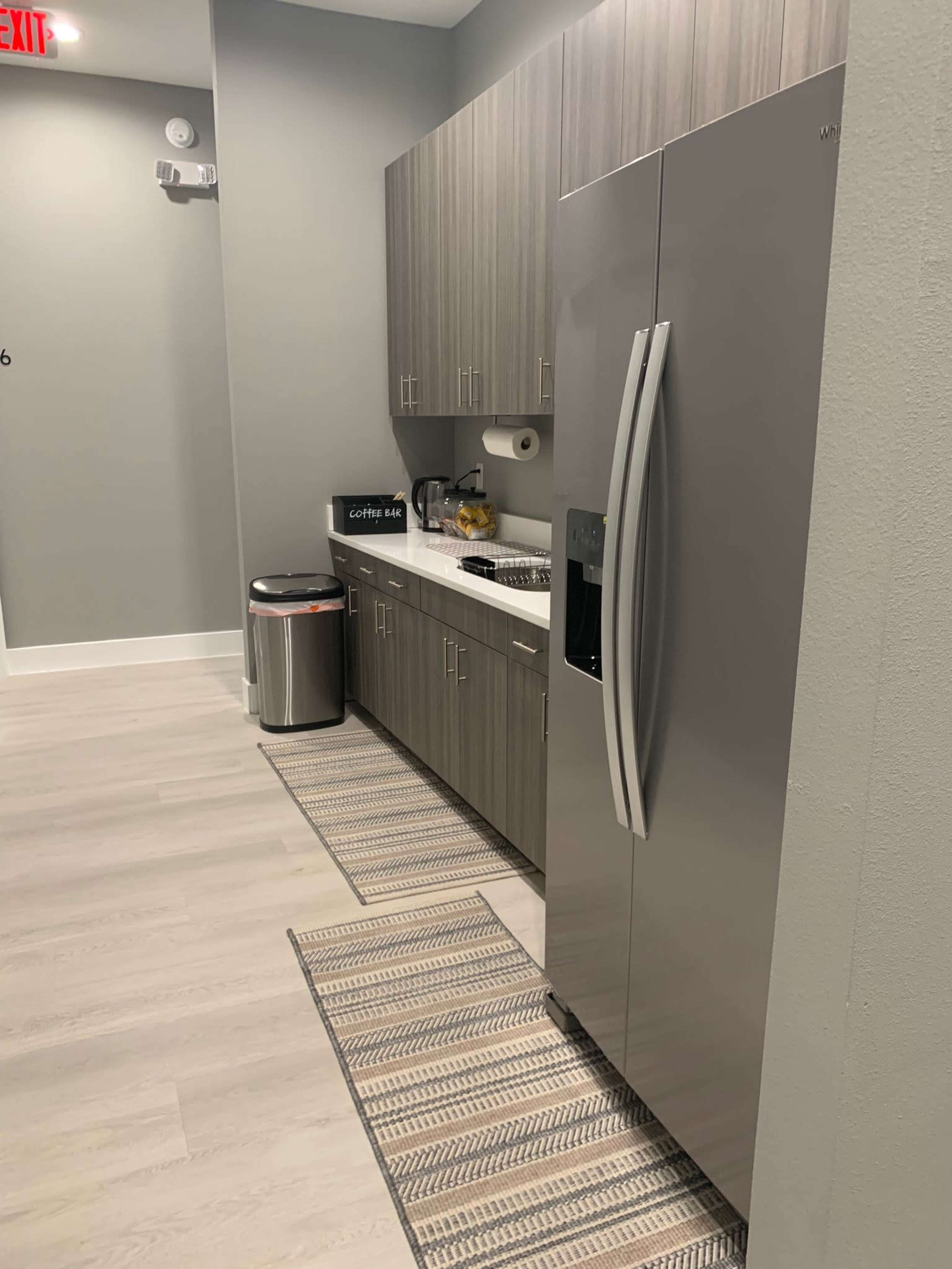 The image shows a modern kitchen featuring a stainless steel refrigerator, wooden cabinets, a countertop with kitchen appliances, and a trash can.