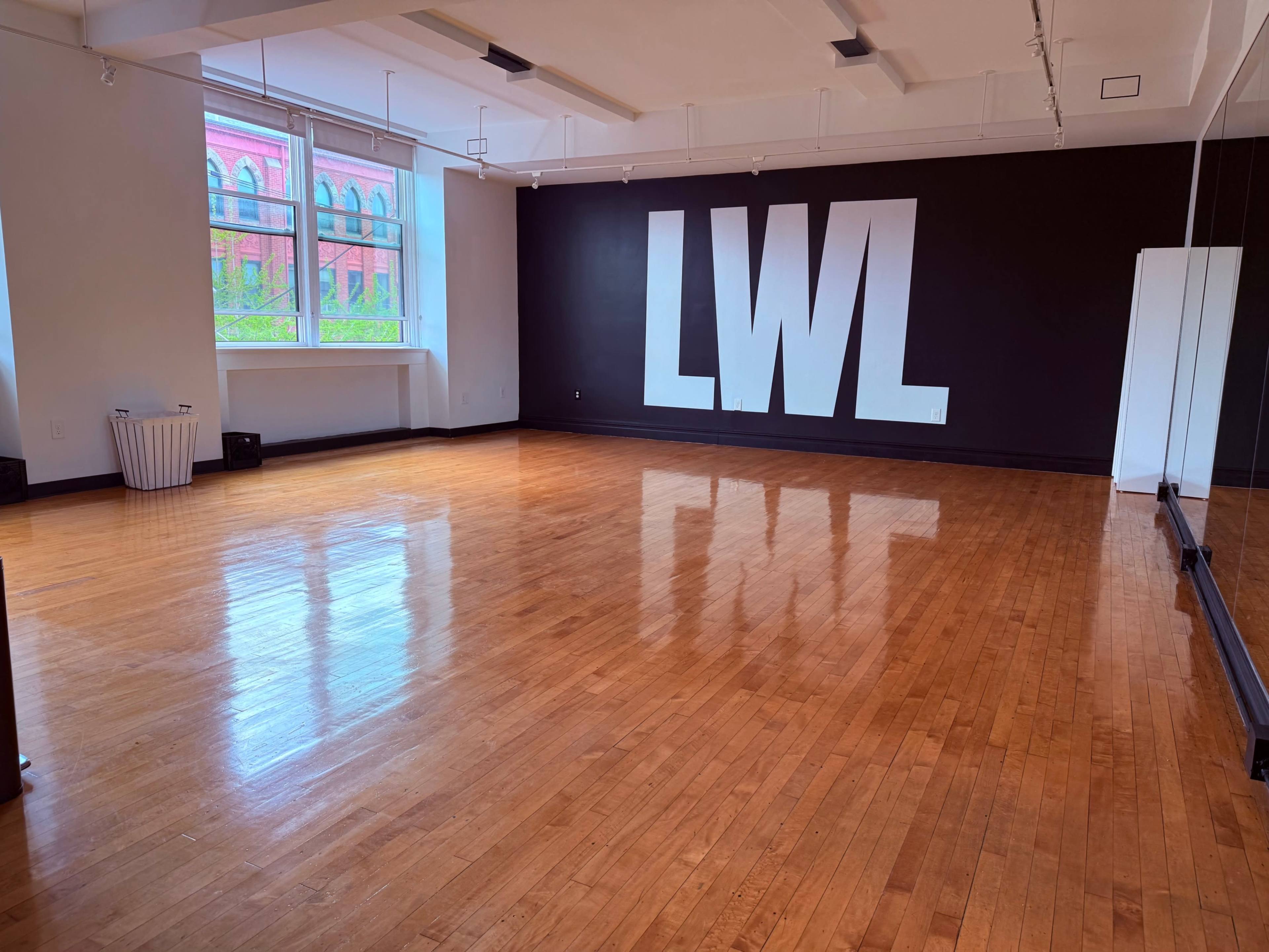 The image shows an empty dance studio with polished wooden floors, a large wall featuring the letters "LWL," and a mirror on one side.