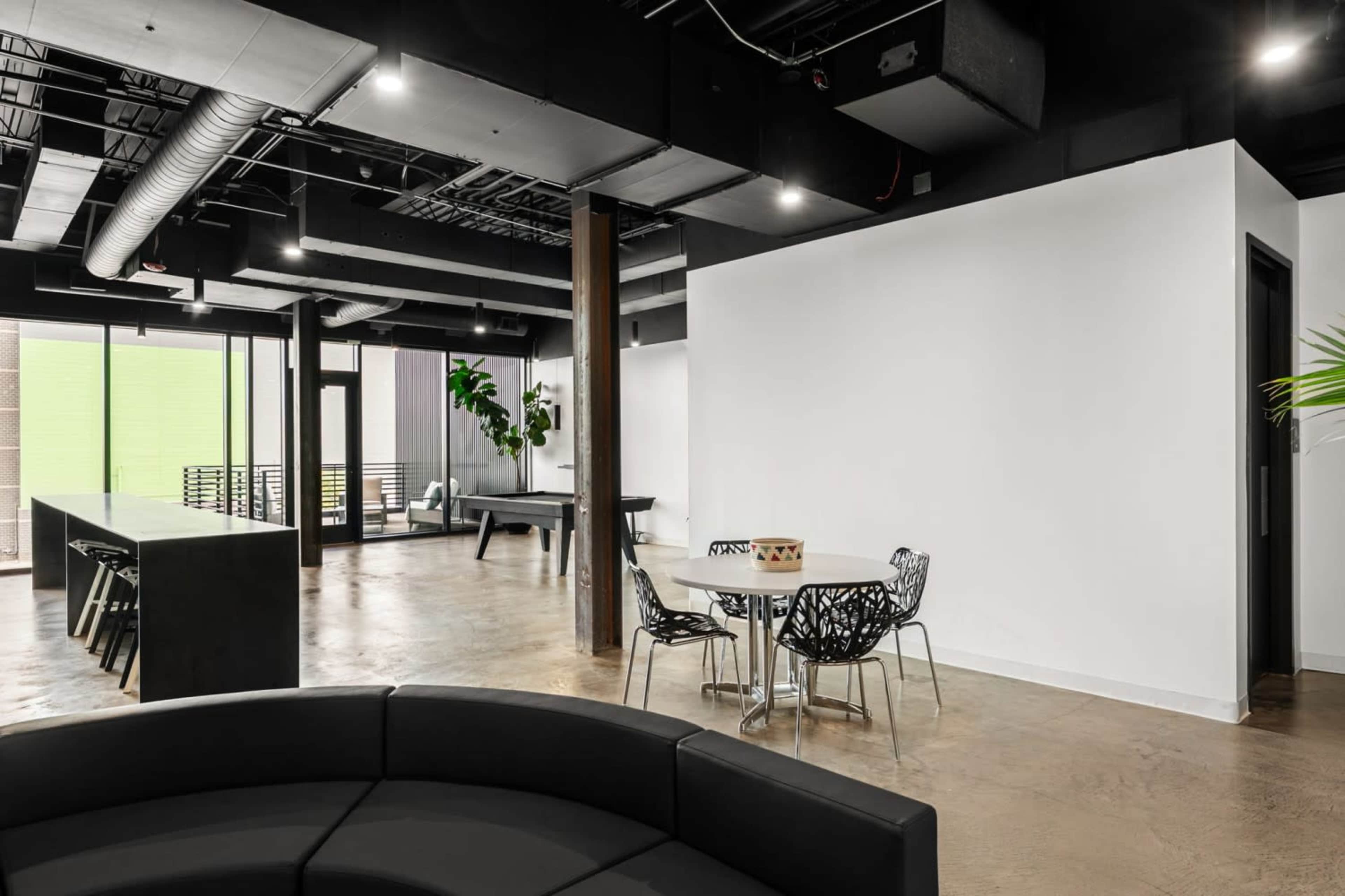 The image shows a modern office space featuring a seating area with a black circular couch, a table with chairs, and large windows providing natural light.