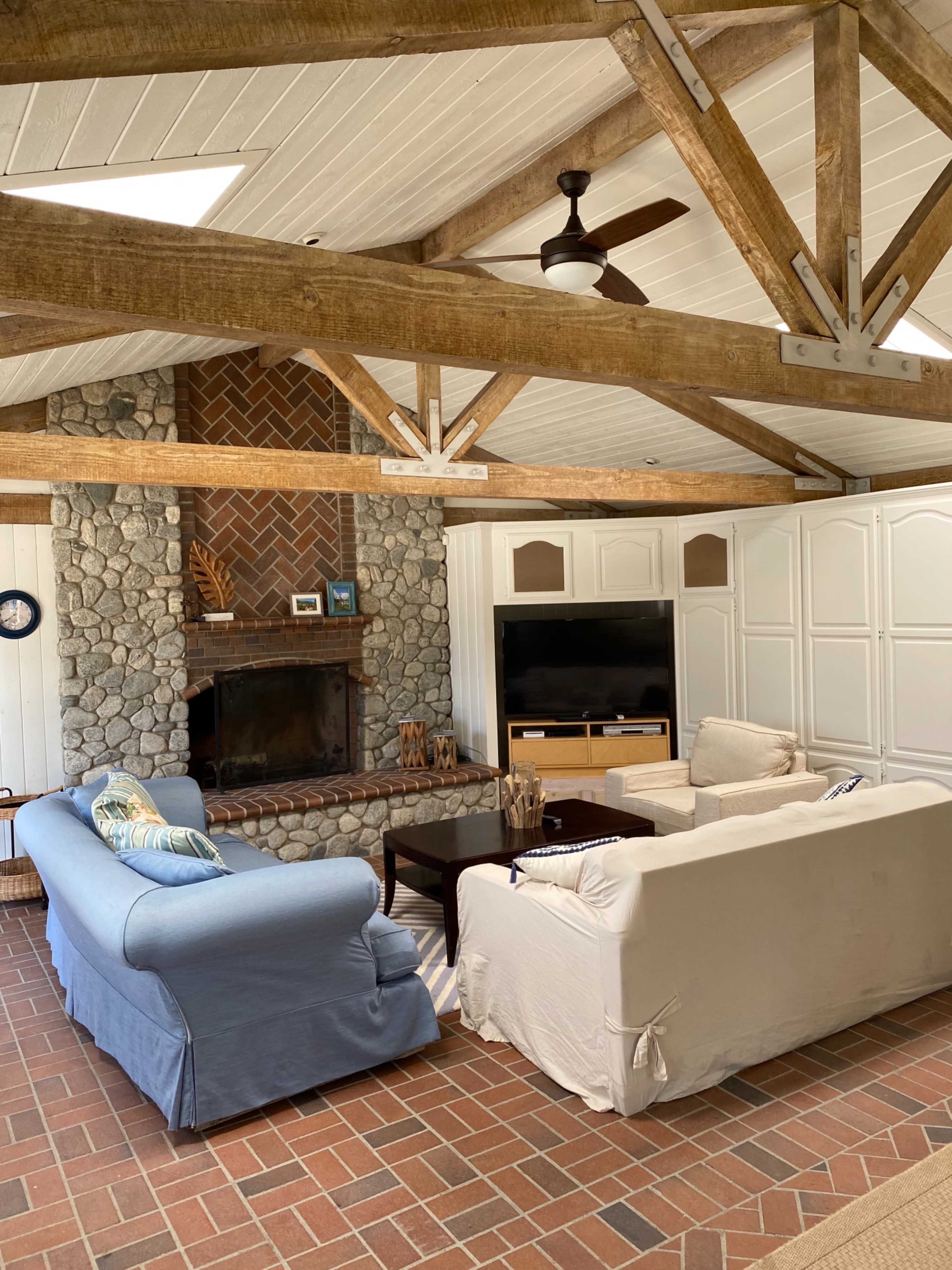 A cozy living room with stone walls, a brick fireplace, and wooden beam ceilings, featuring two sofas and a television.