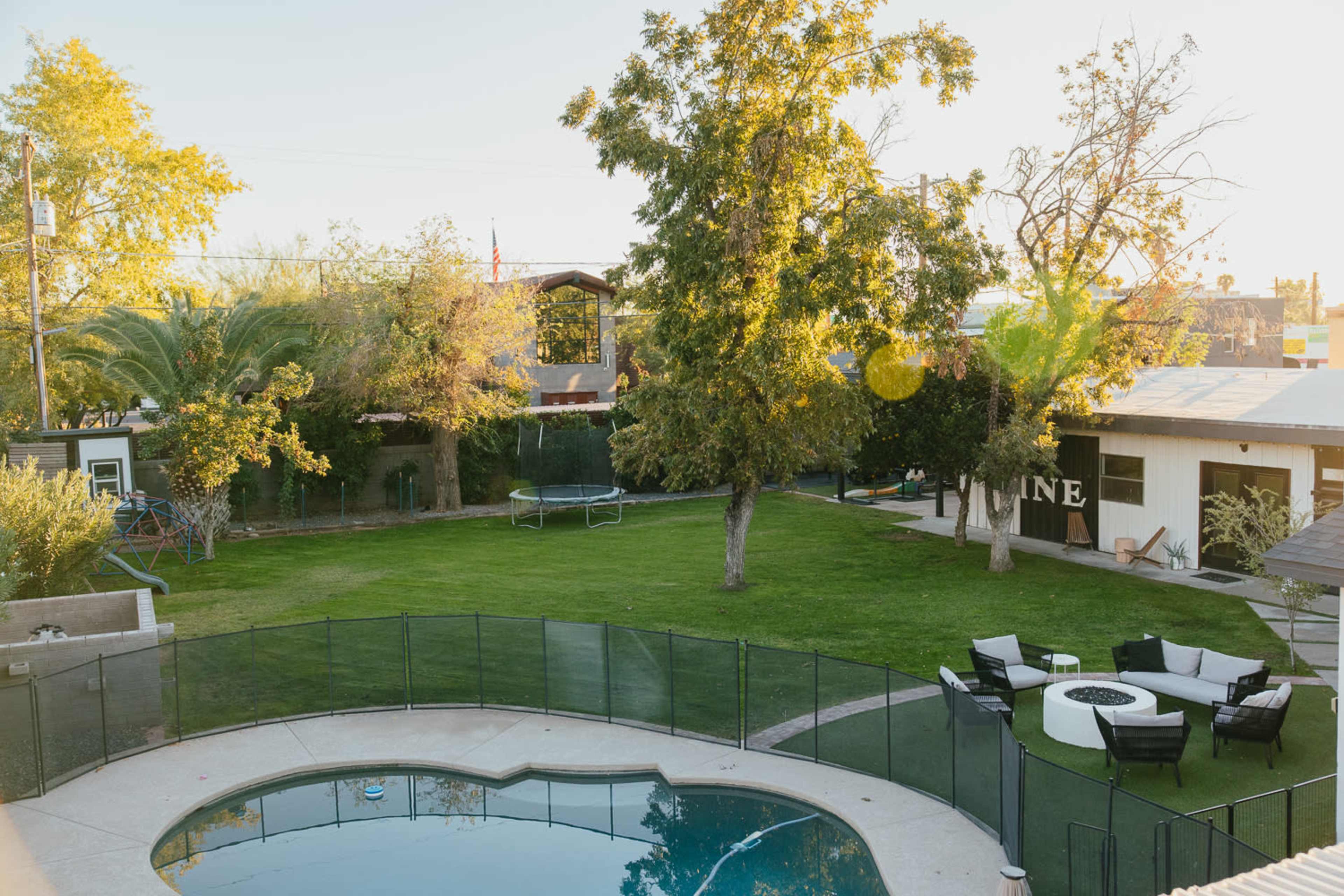 A backyard features a swimming pool surrounded by a safety fence, with a grassy area, several trees, and outdoor seating arrangements.