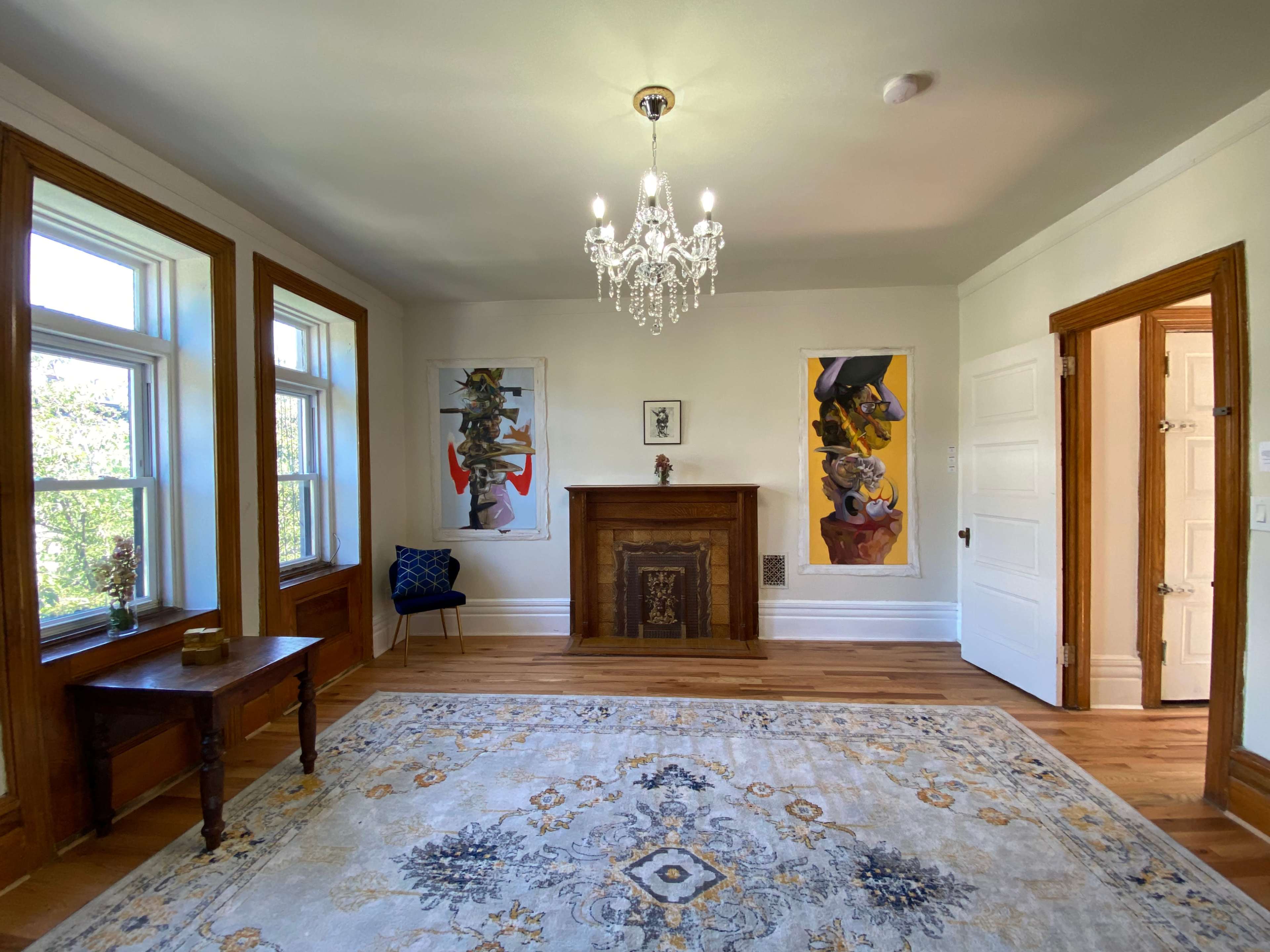 The room features a chandelier above a decorative fireplace, flanked by large windows and two pieces of artwork on the walls.