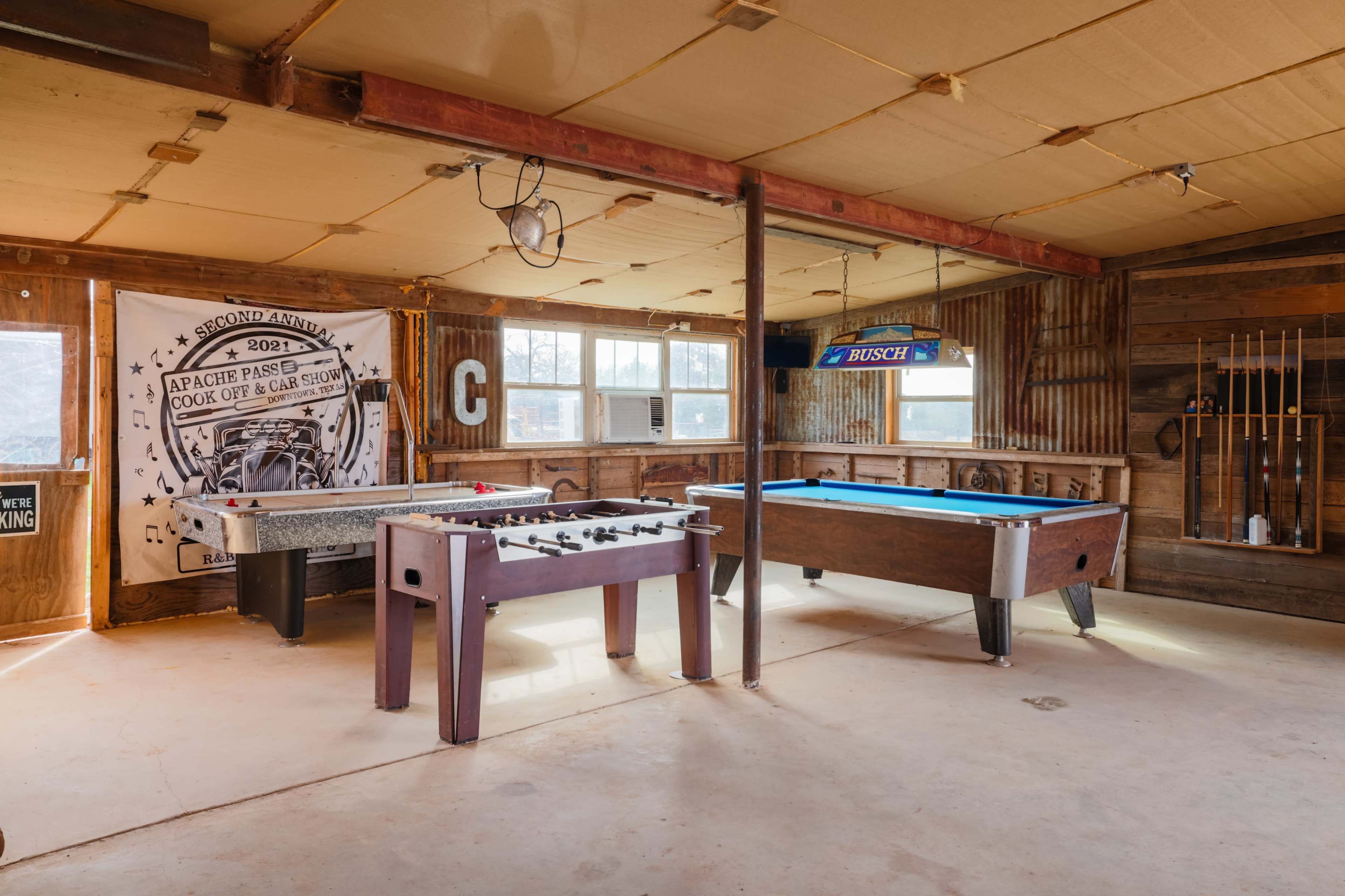 The image shows a rustic game room with a foosball table, a pool table, and a large wall banner decorated with a vintage design.
