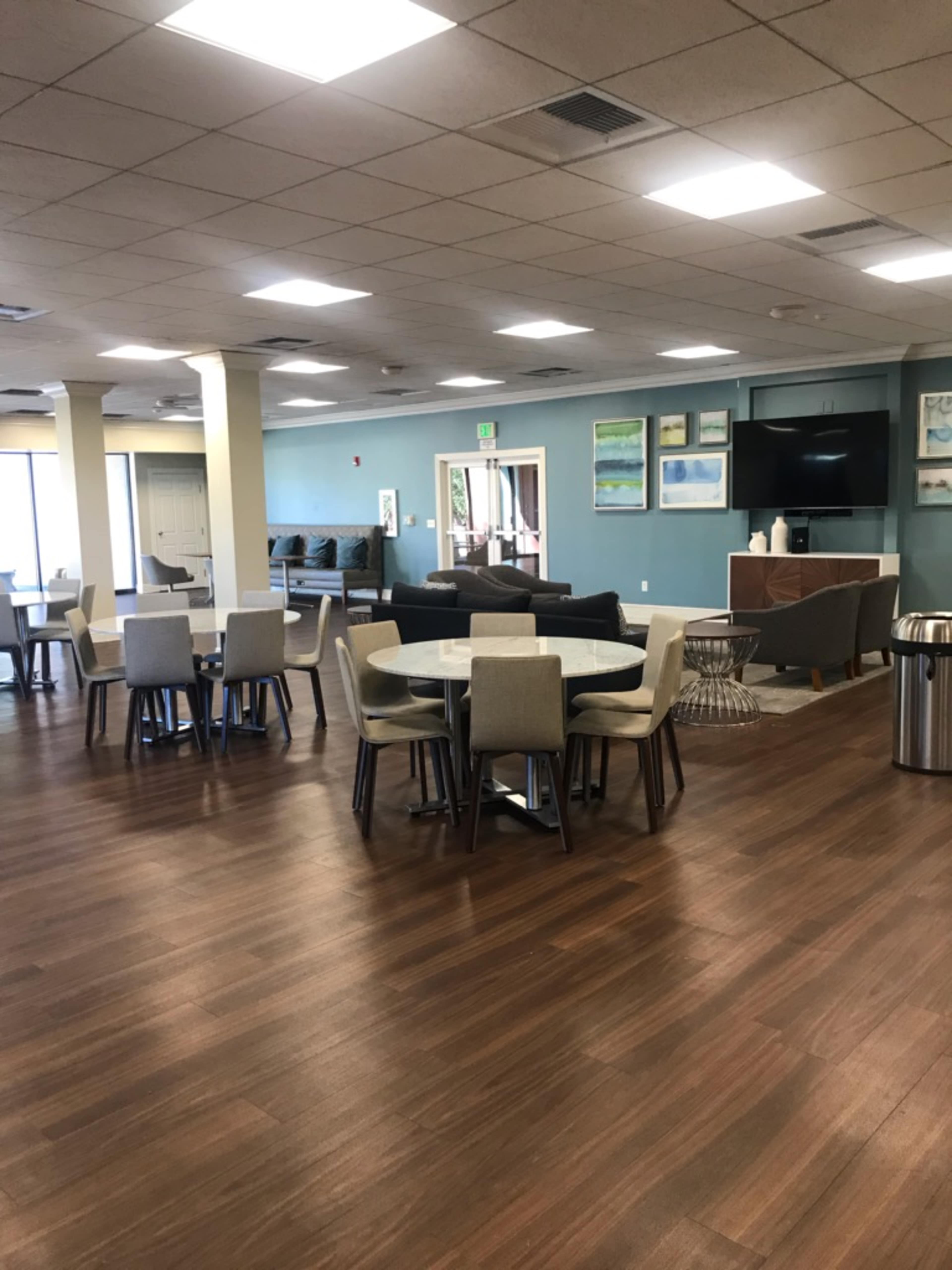 A spacious communal area featuring several round tables with chairs, a seating area with sofas, and a kitchenette against a light blue wall.