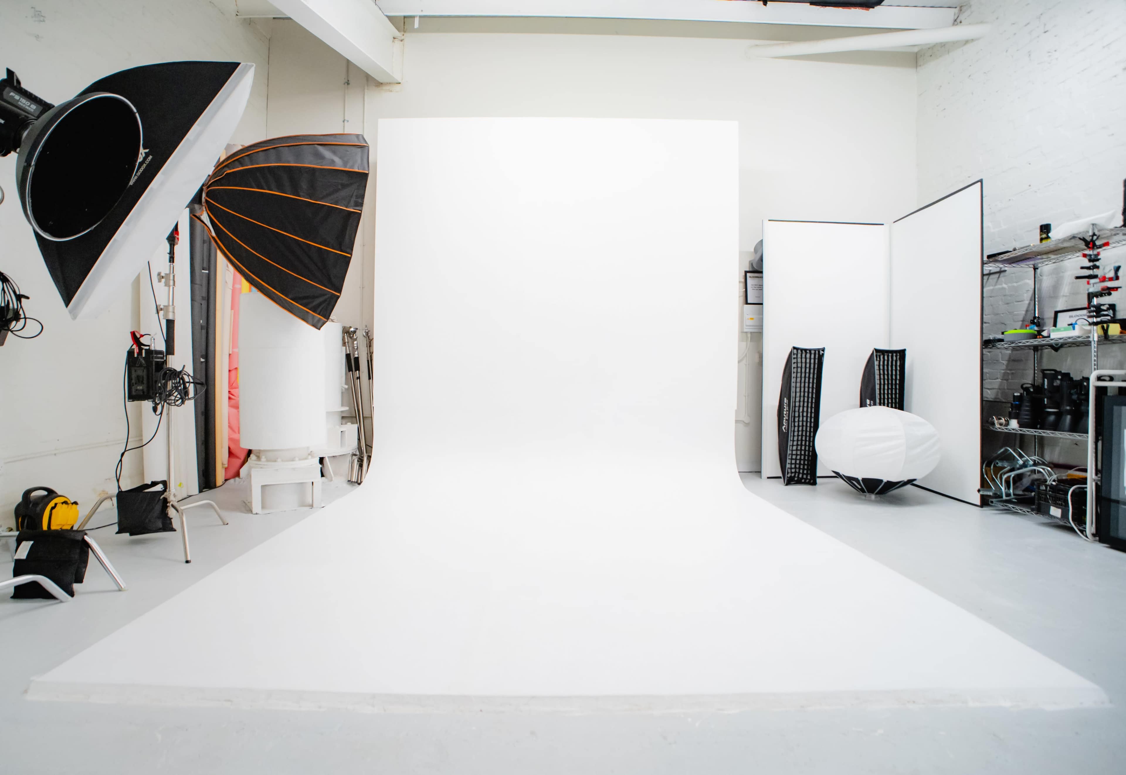 The image shows a photography studio with a large white seamless backdrop, lighting equipment, and various props arranged along the walls.