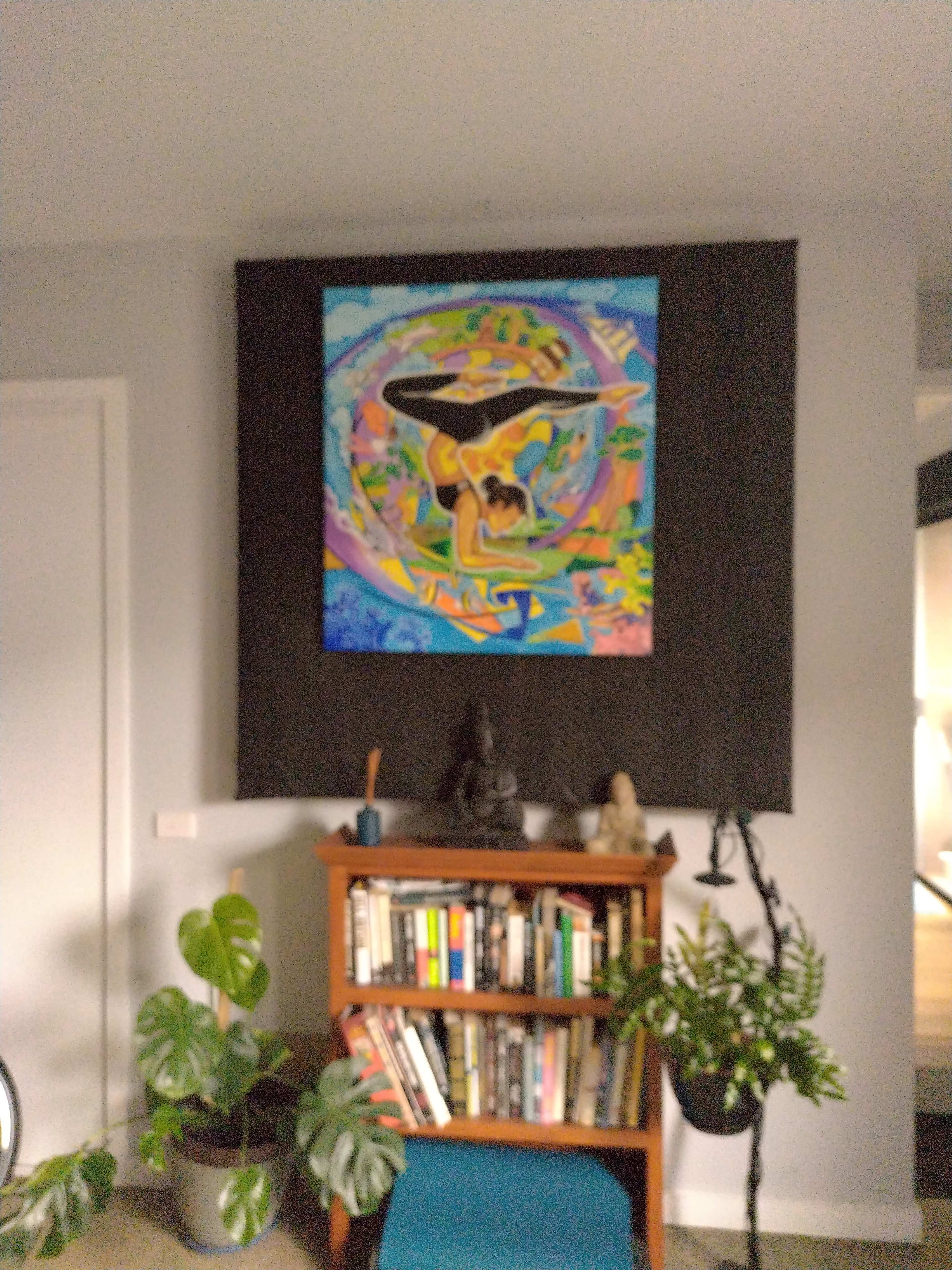 The image shows a colorful painting hung on a wall above a wooden bookshelf filled with books, alongside indoor plants and decorative figurines.