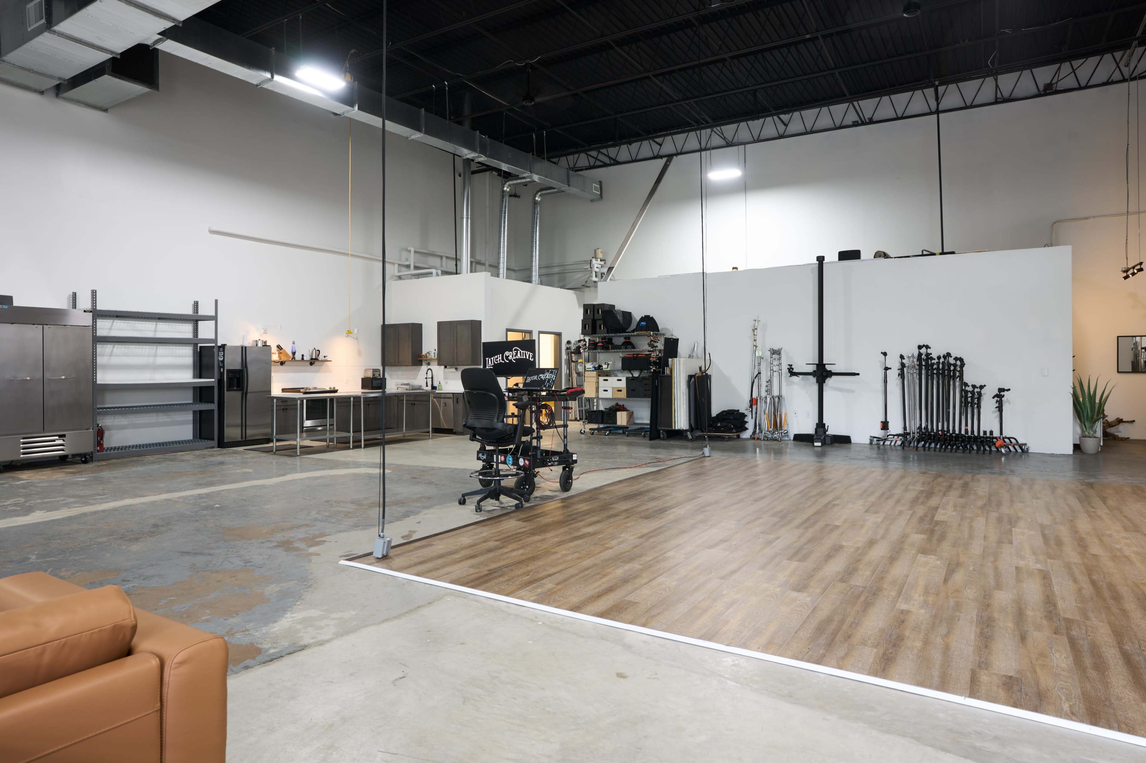 The image shows a spacious, partially furnished studio featuring a kitchen area, a seating arrangement, and various fitness equipment arranged against the wall.