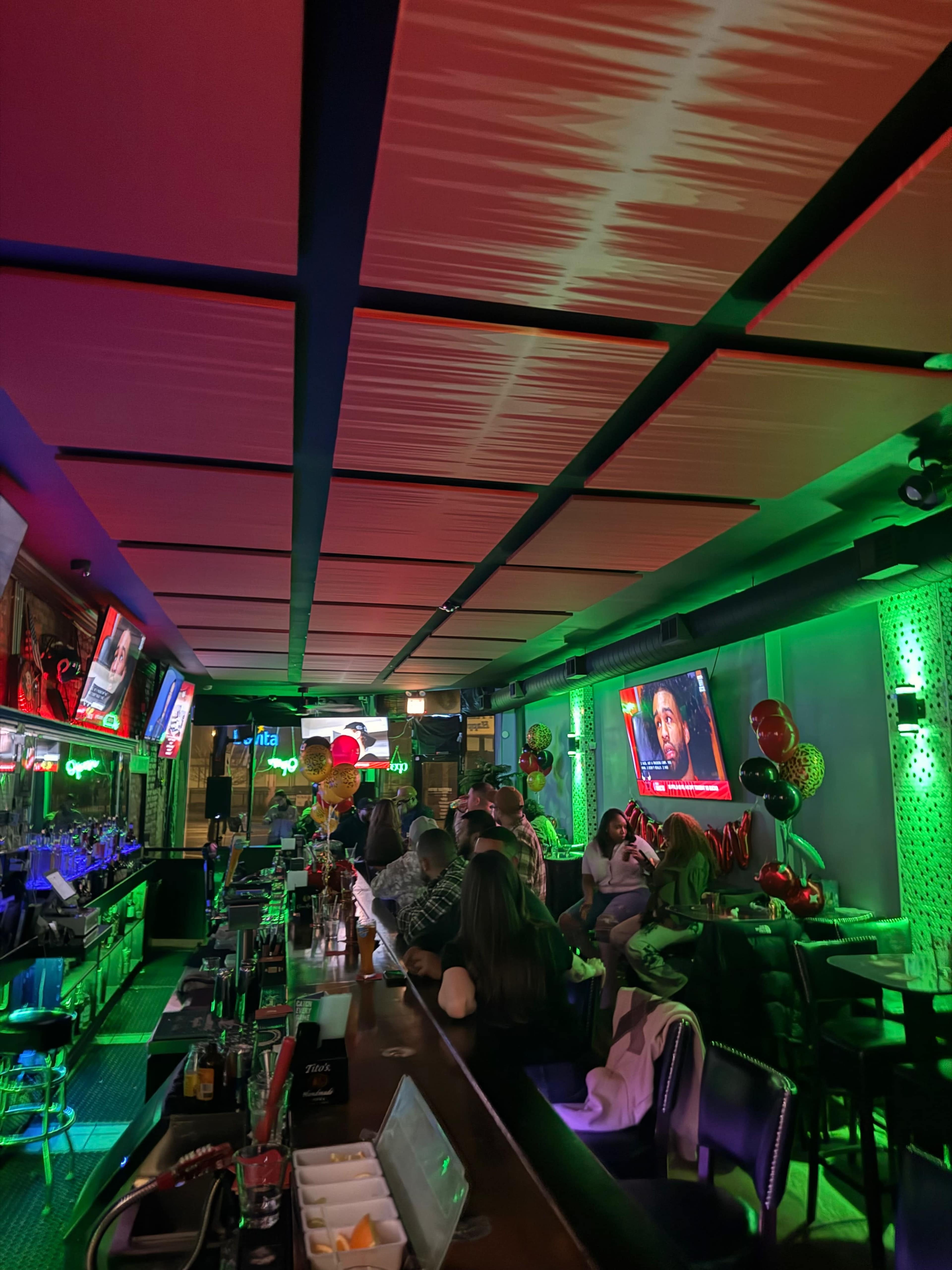 The image shows a dimly lit bar with a long counter, tables occupied by patrons, and colorful lighting accentuating the decor.