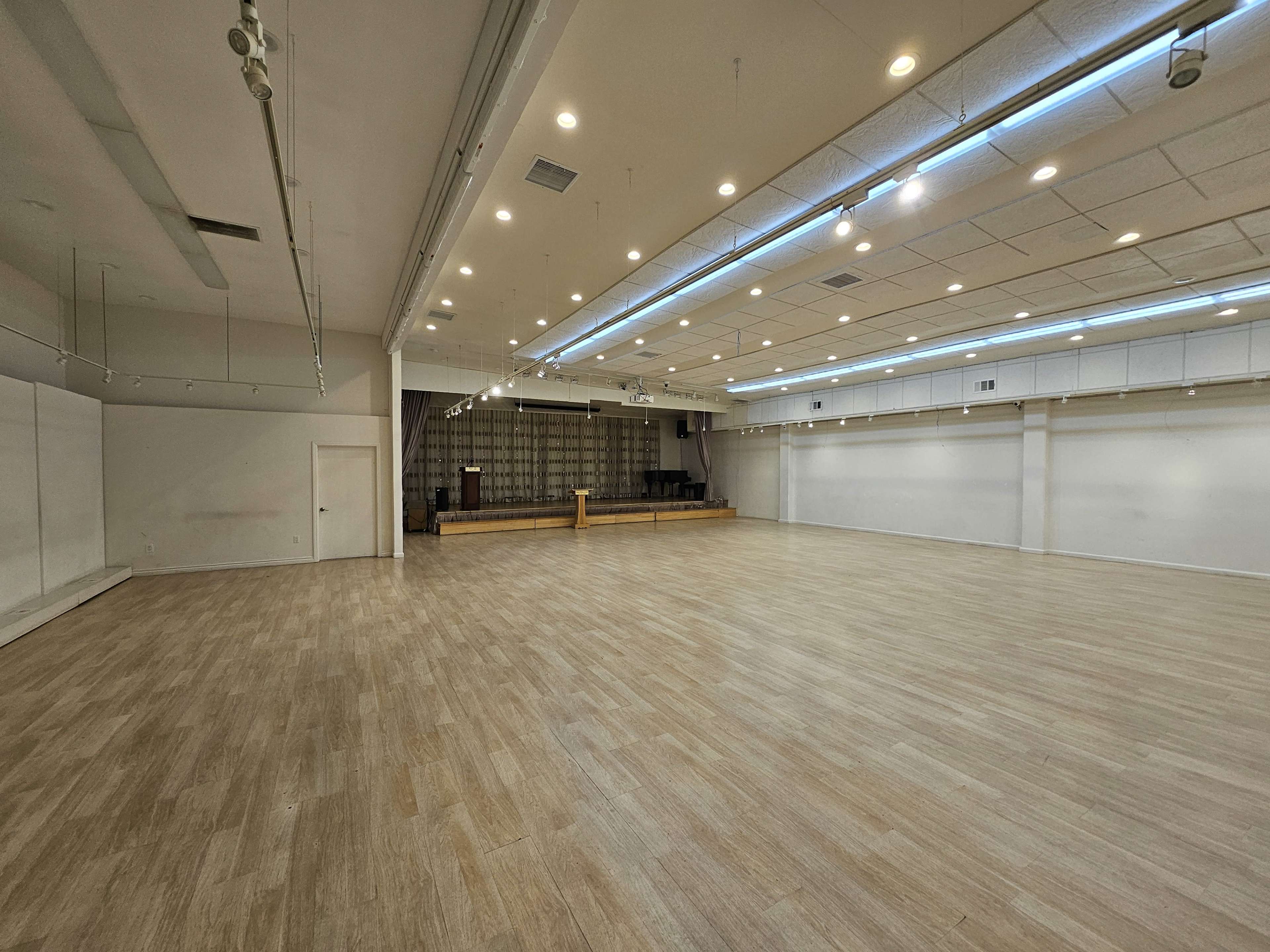 A spacious, empty indoor venue with wooden floors, white walls, and a stage at the far end.