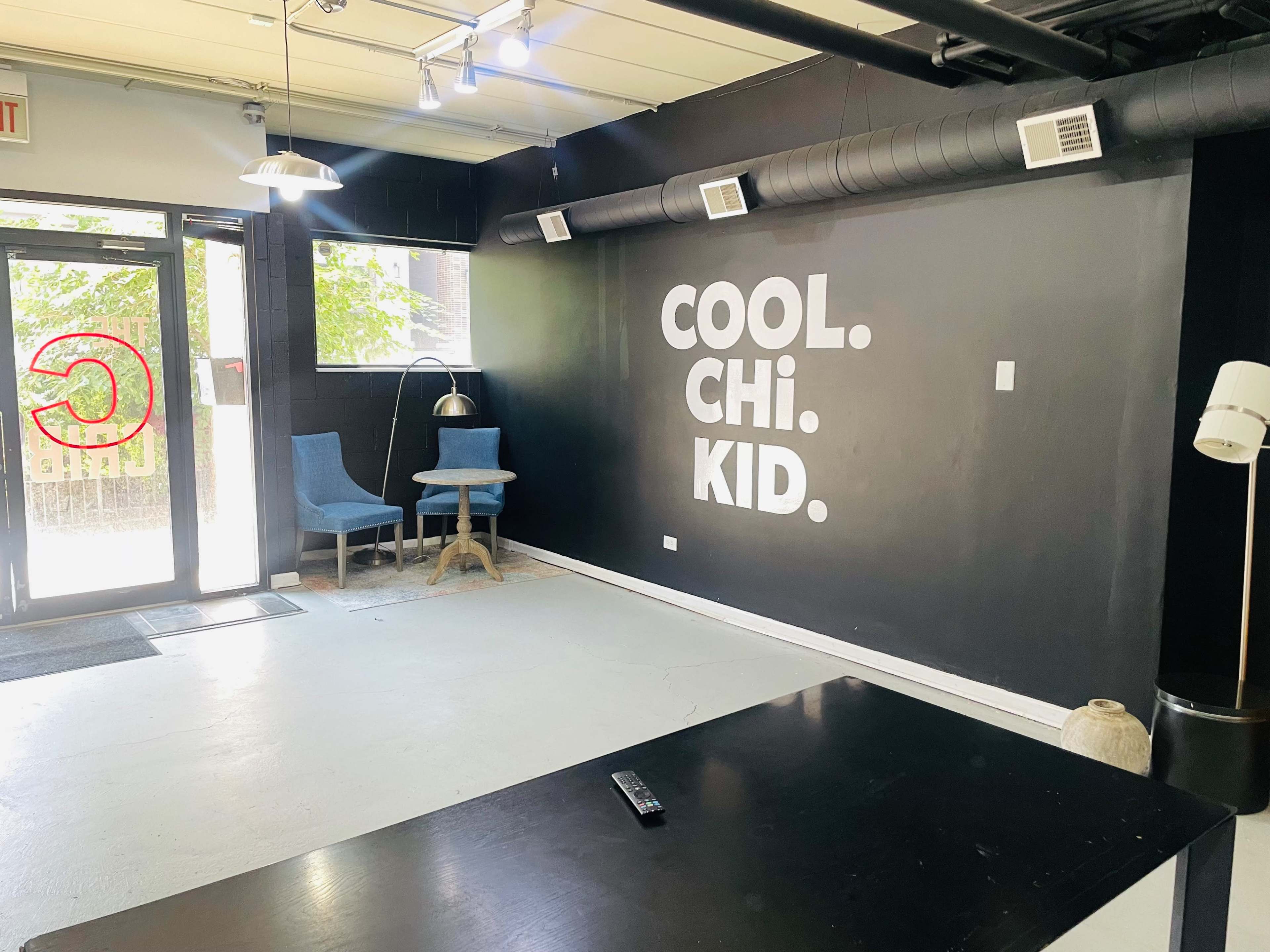 The interior of a room features a black wall with the words "COOL. CHI. KID." prominently displayed, two blue chairs, a small table, and a door leading outside.