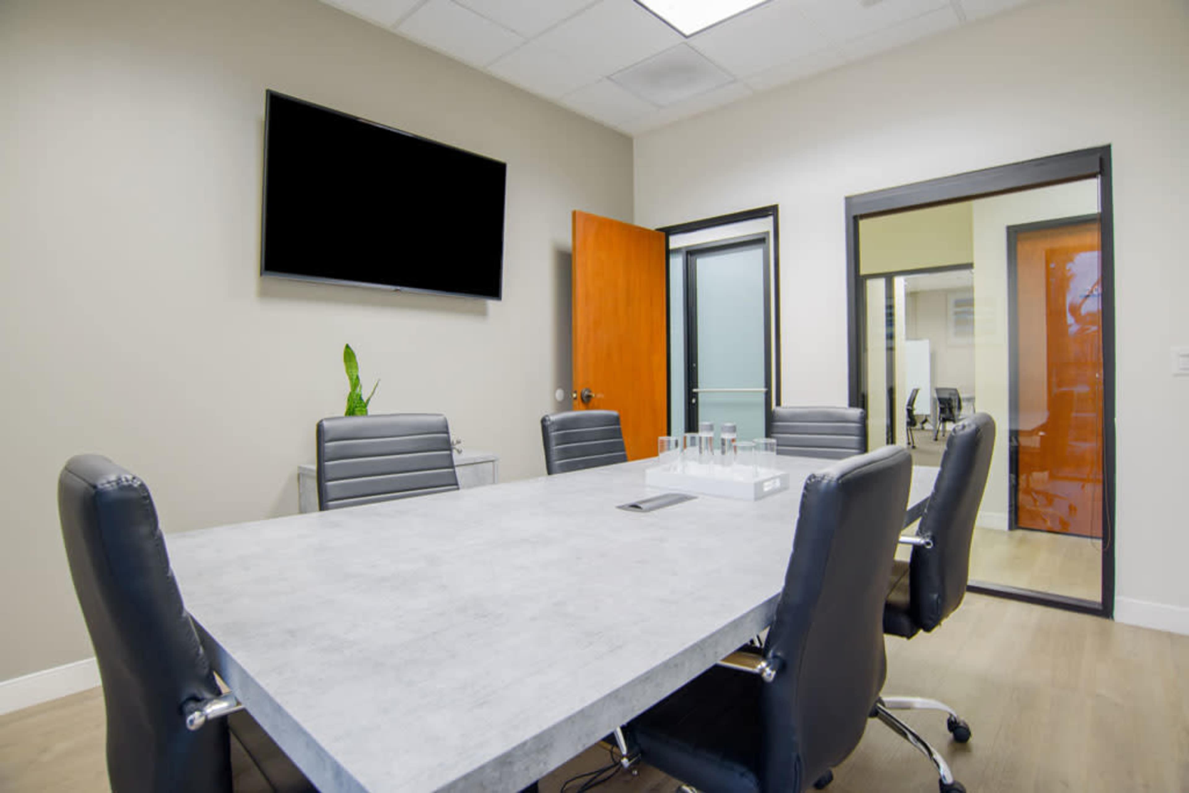 A conference room features a large gray table surrounded by black leather chairs, with a television mounted on the wall and a glass door leading to another office.
