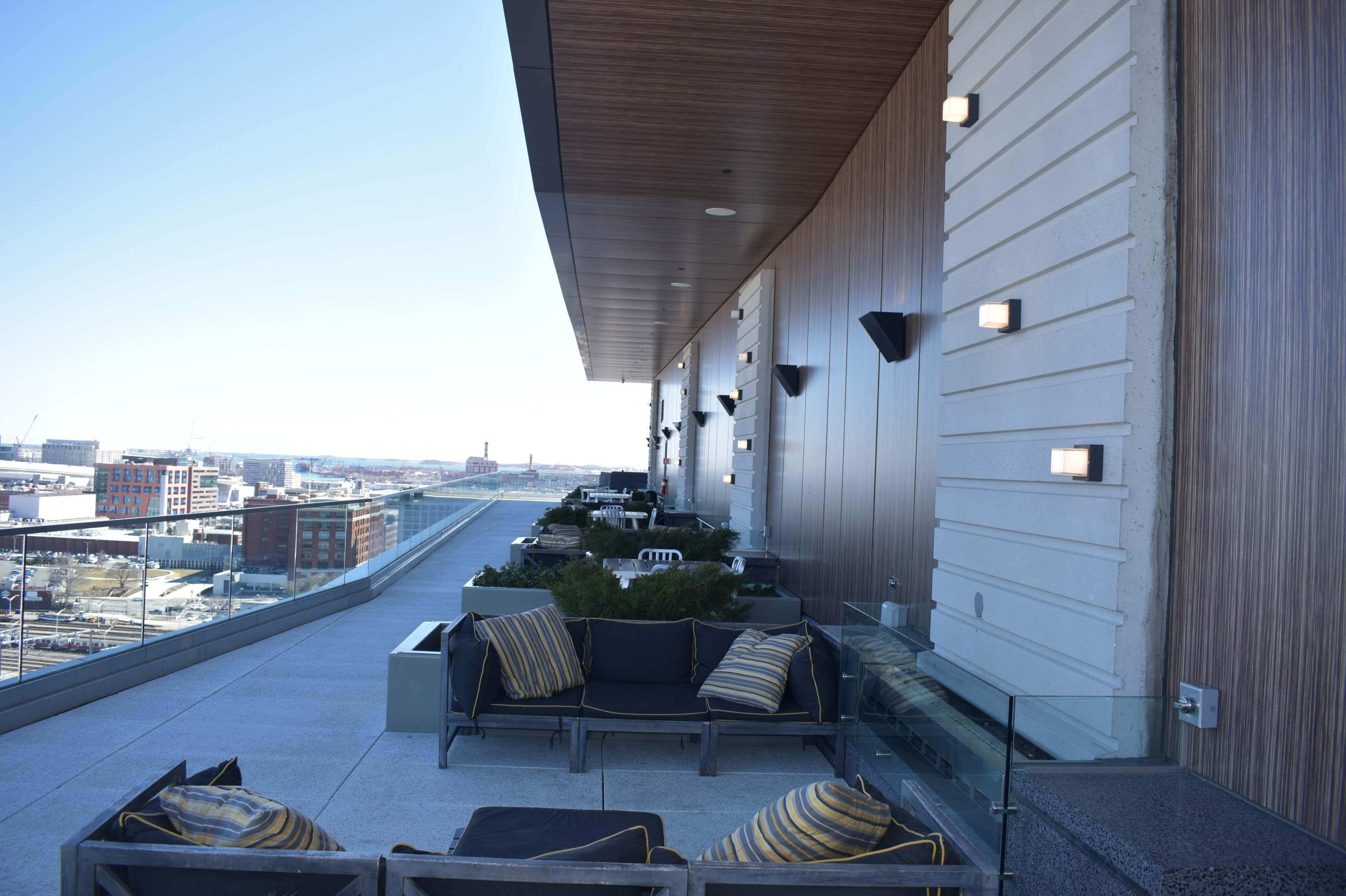 A modern rooftop terrace with seating areas, planters, and wall-mounted lights, overlooking a city skyline.