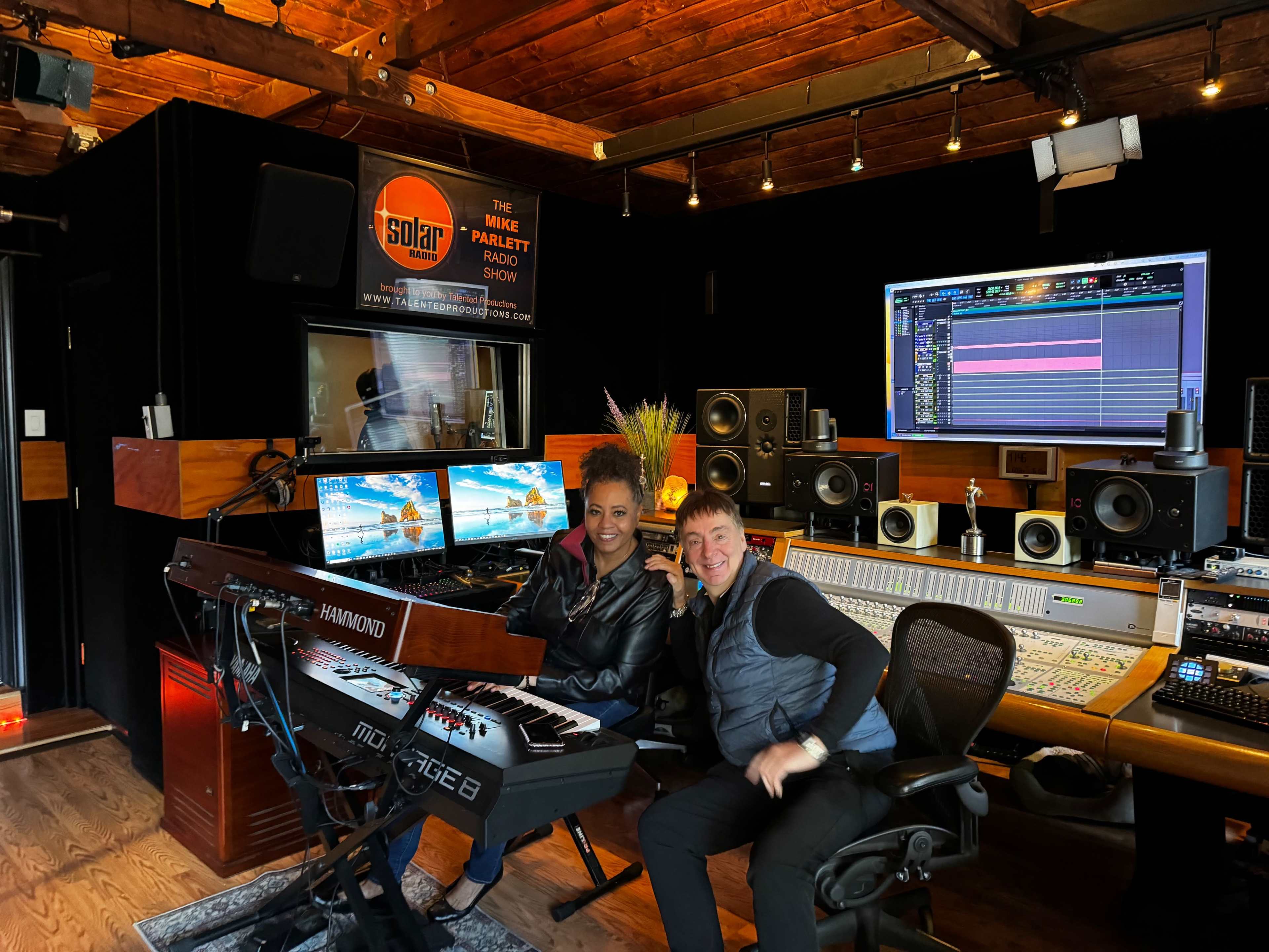 Two people are sitting in a music recording studio, surrounded by equipment and monitors displaying audio tracks.