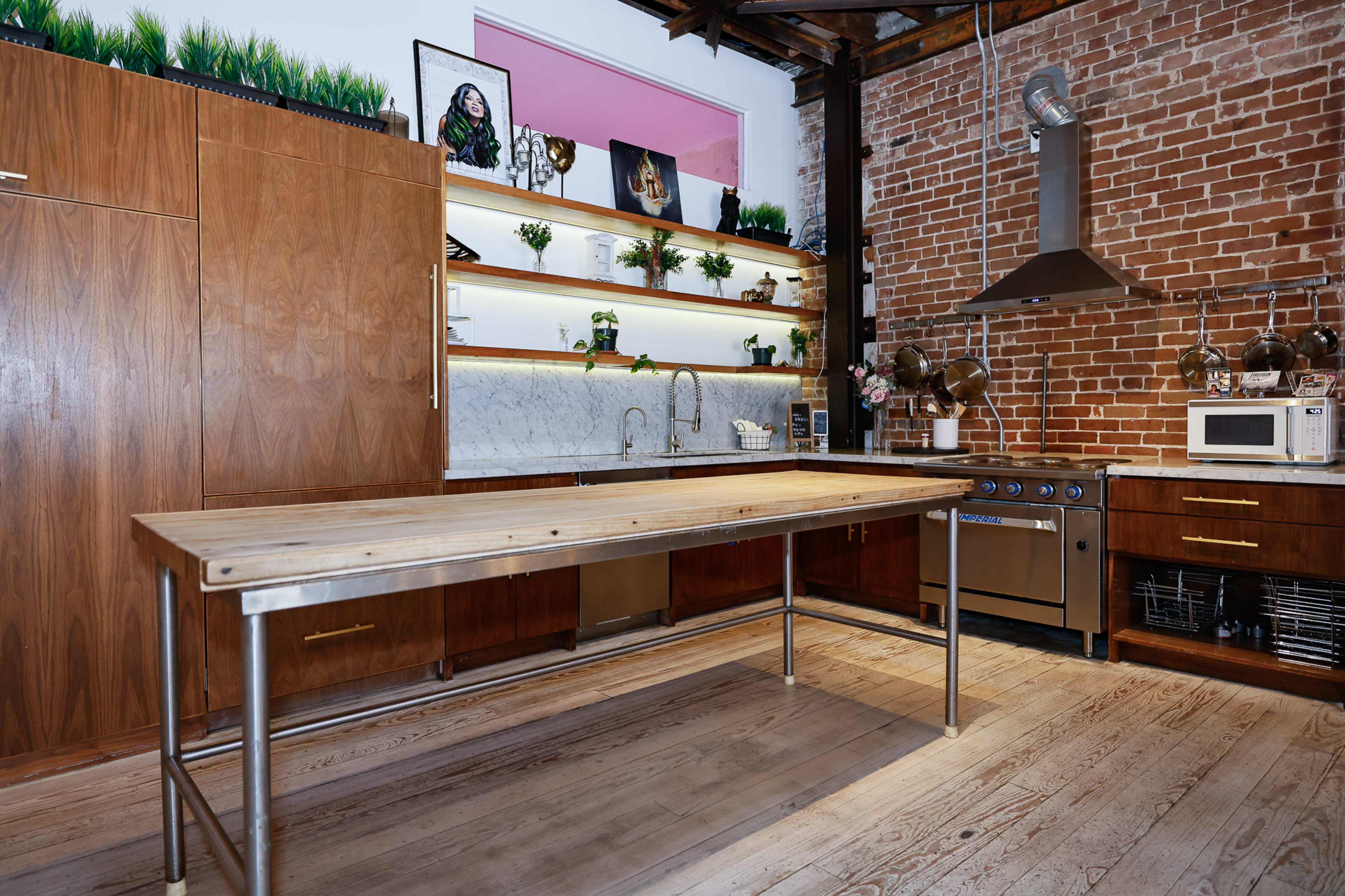 The image features a modern kitchen with a long wooden table, stainless steel appliances, and exposed brick walls, illuminated by shelves of plants and decorative items.