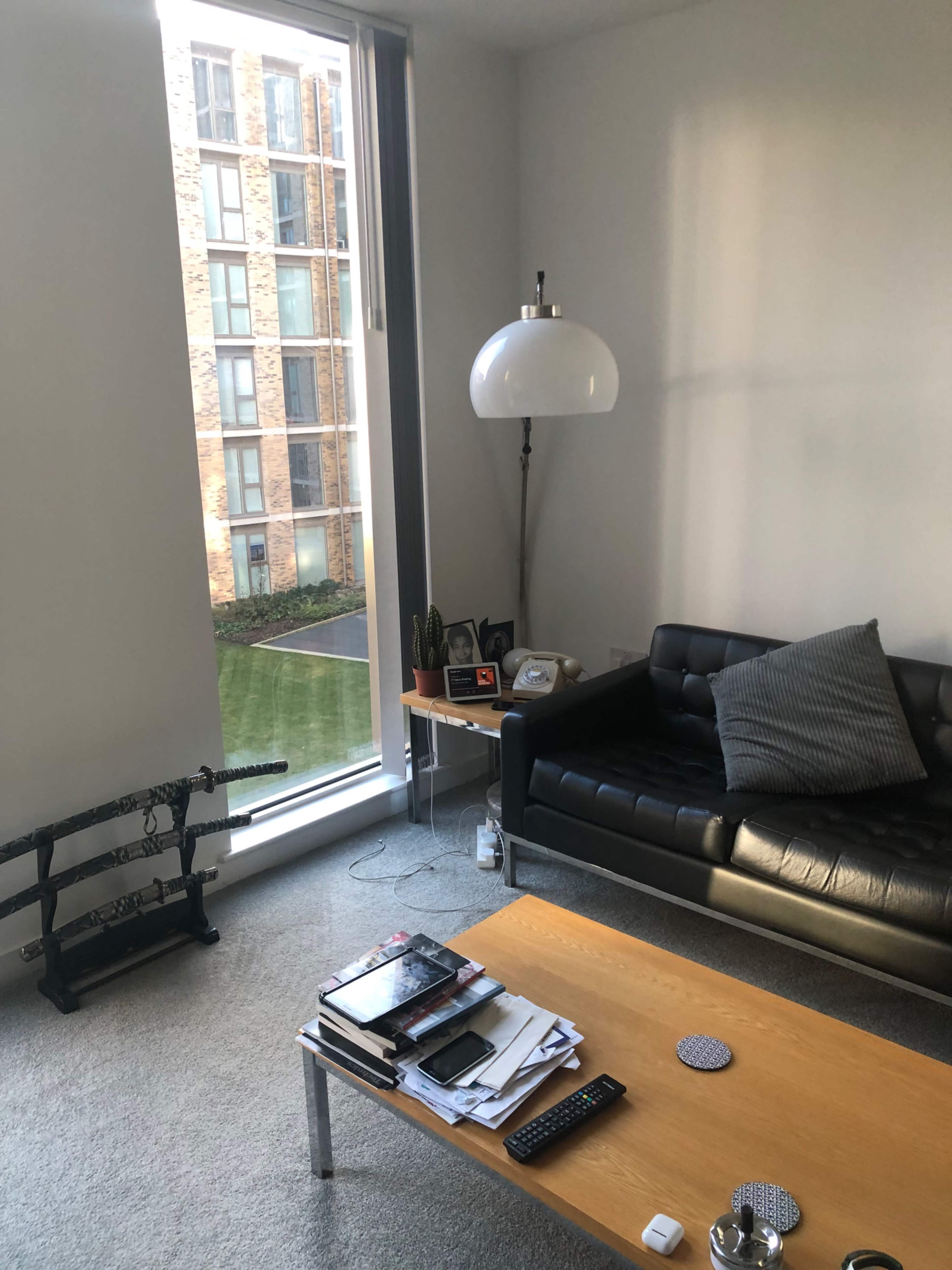 The image shows a modern living room with a black sofa, a wooden coffee table cluttered with items, and a large window revealing a view of a building adjacent to the room.