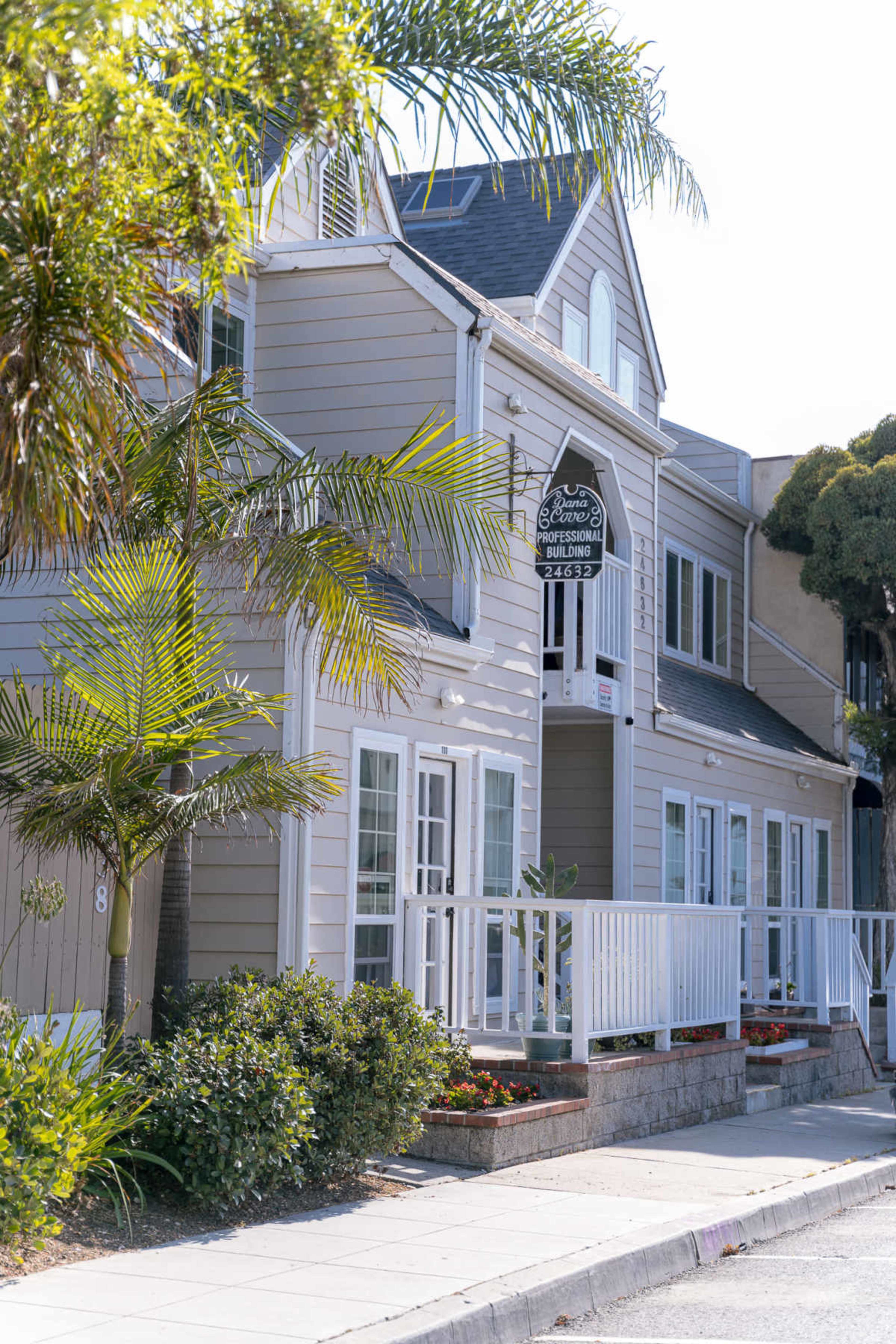 The image shows a well-maintained two-story building with a white exterior, palm trees, and a sign overhead, located in a sunny urban area.