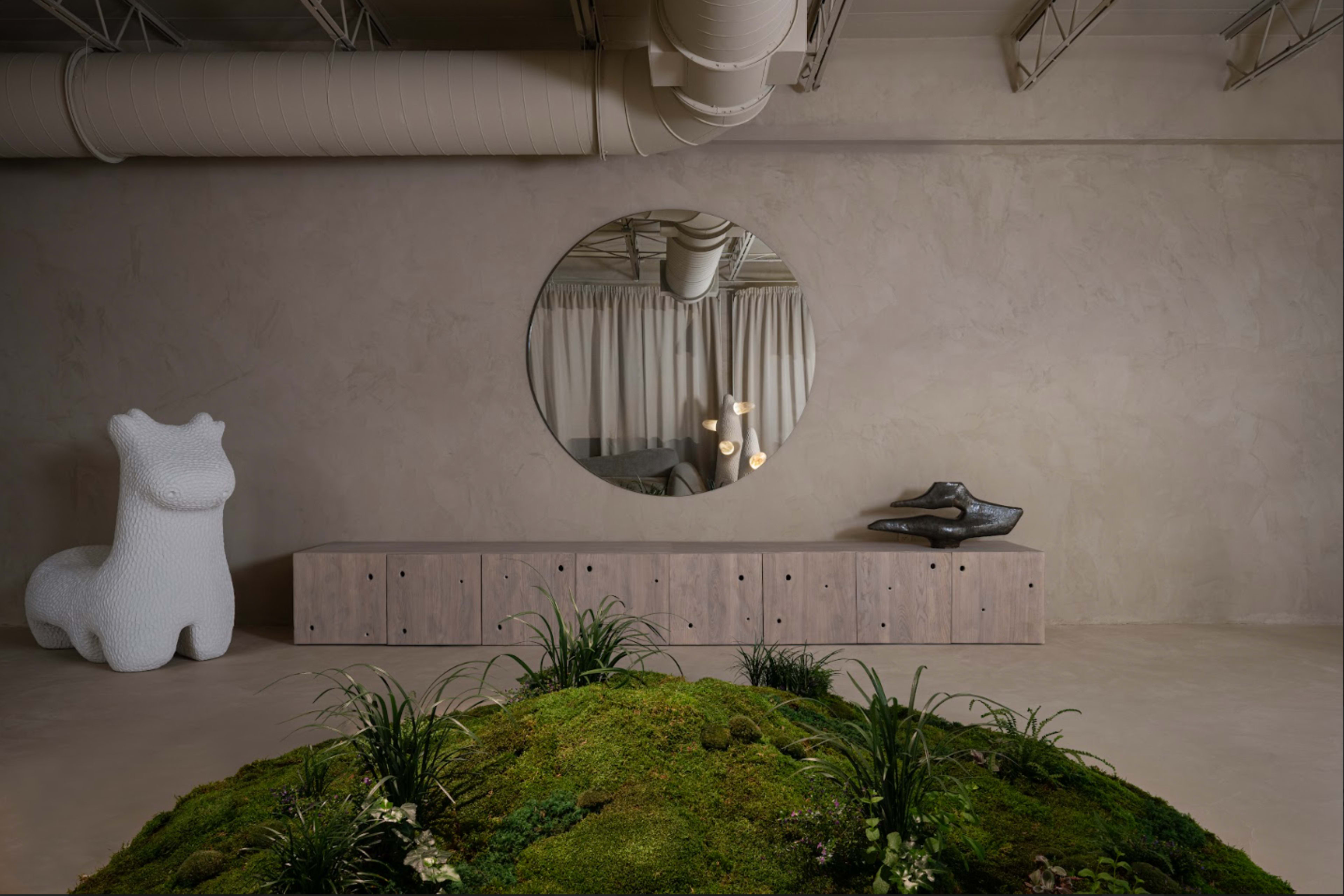The image depicts a minimalist interior space featuring a round mirror, a textured storage unit, and a small indoor landscape with moss and plants.