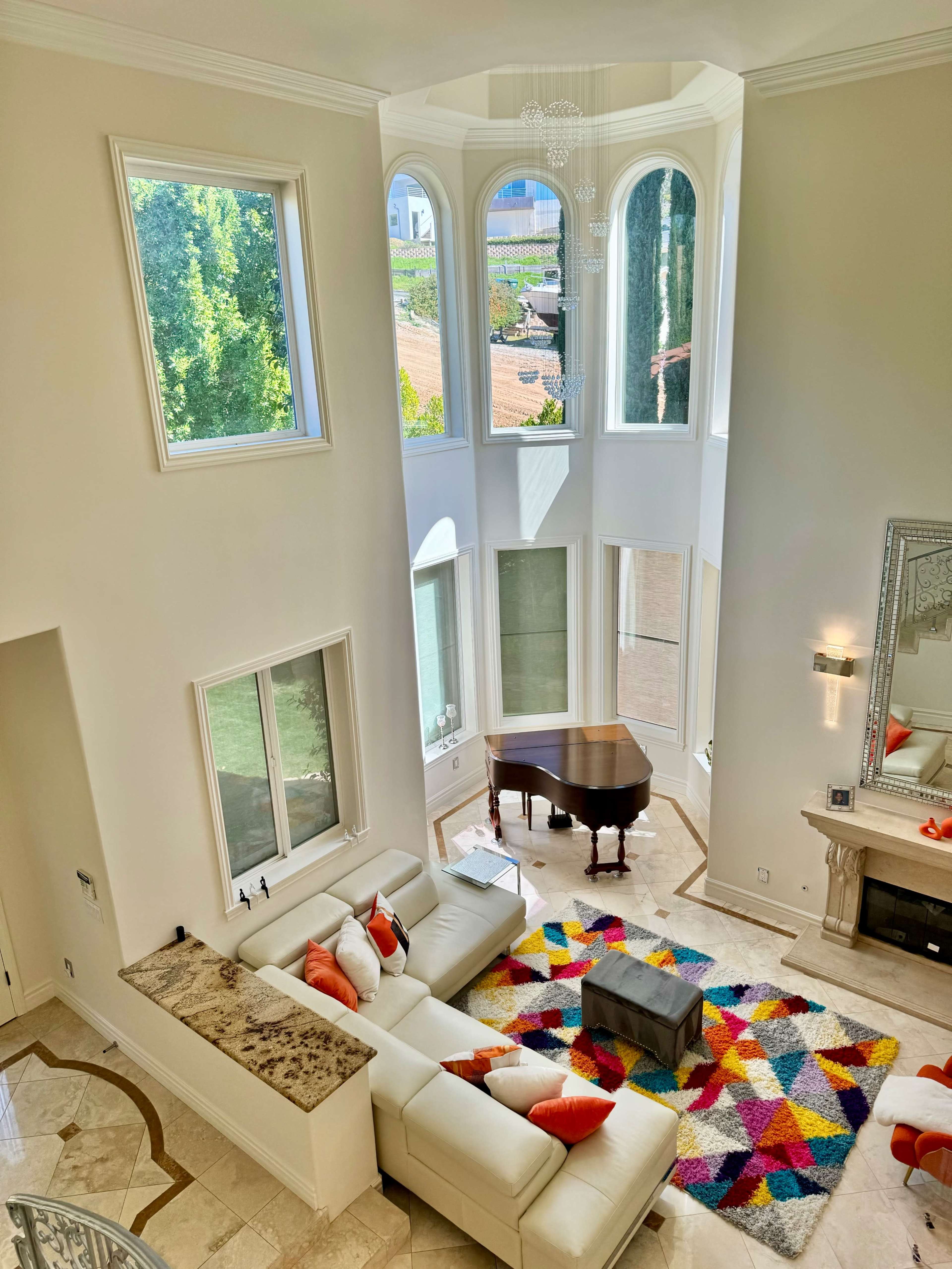 The image shows a spacious living room with a white sectional sofa, colorful rug, a grand piano, and large windows that let in natural light.