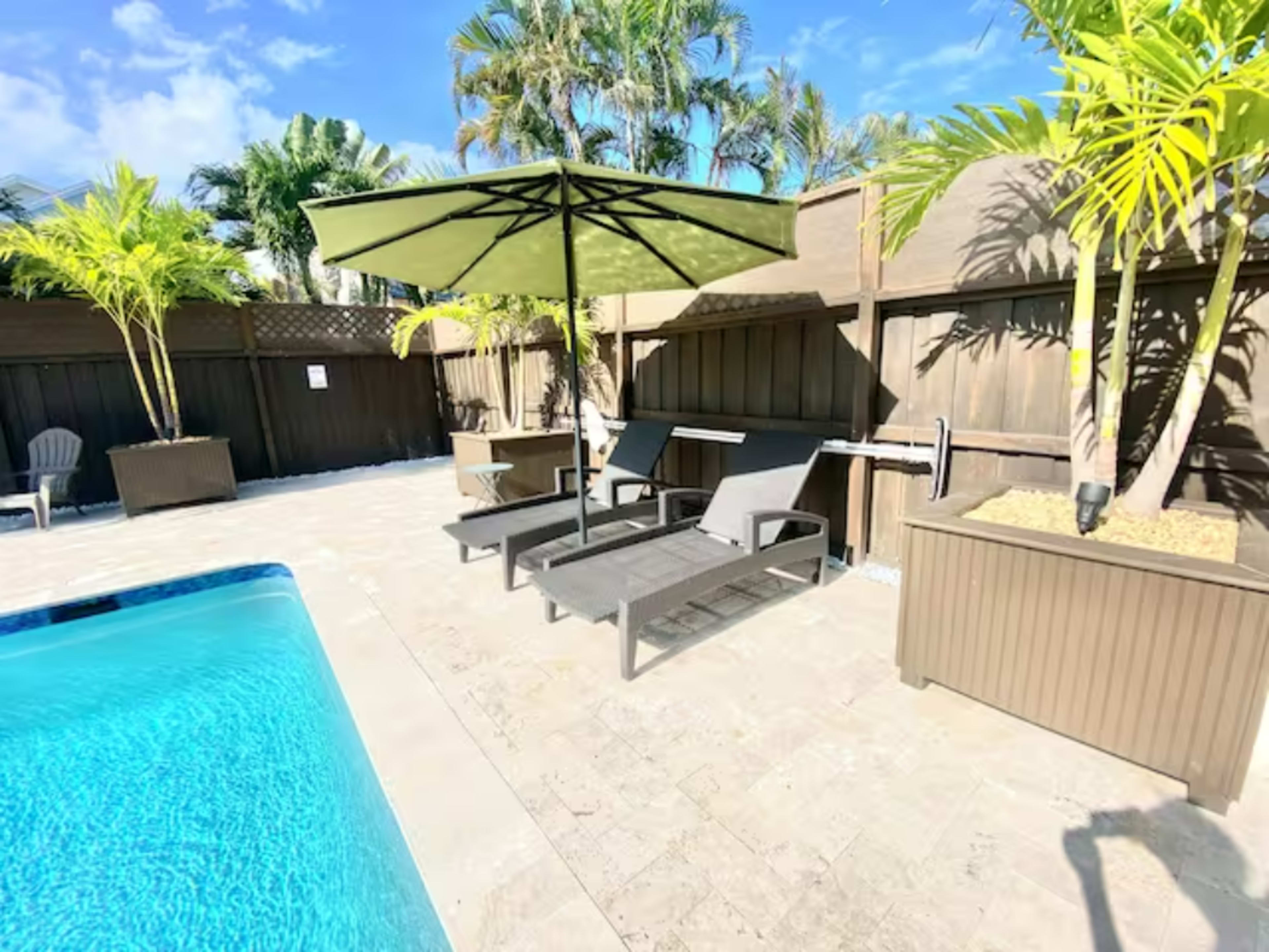 The scene features a swimming pool area with two lounge chairs and an umbrella, surrounded by palm trees and a wooden fence.
