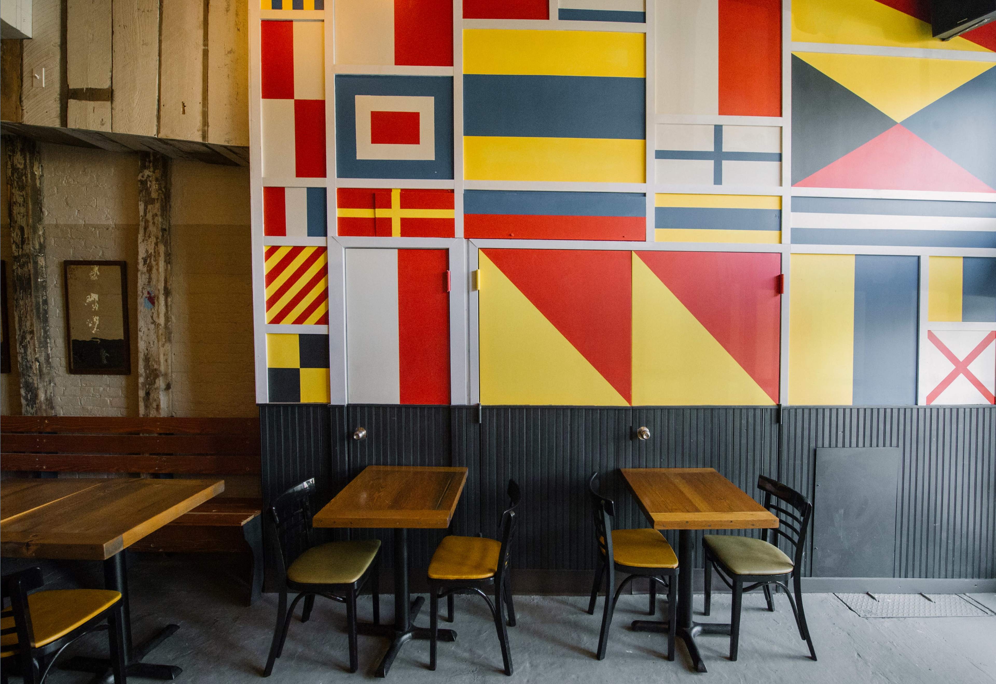 The image shows a restaurant interior with a wall decorated in a vibrant, geometric pattern resembling various flags, and several wooden tables with black chairs arranged in front of it.