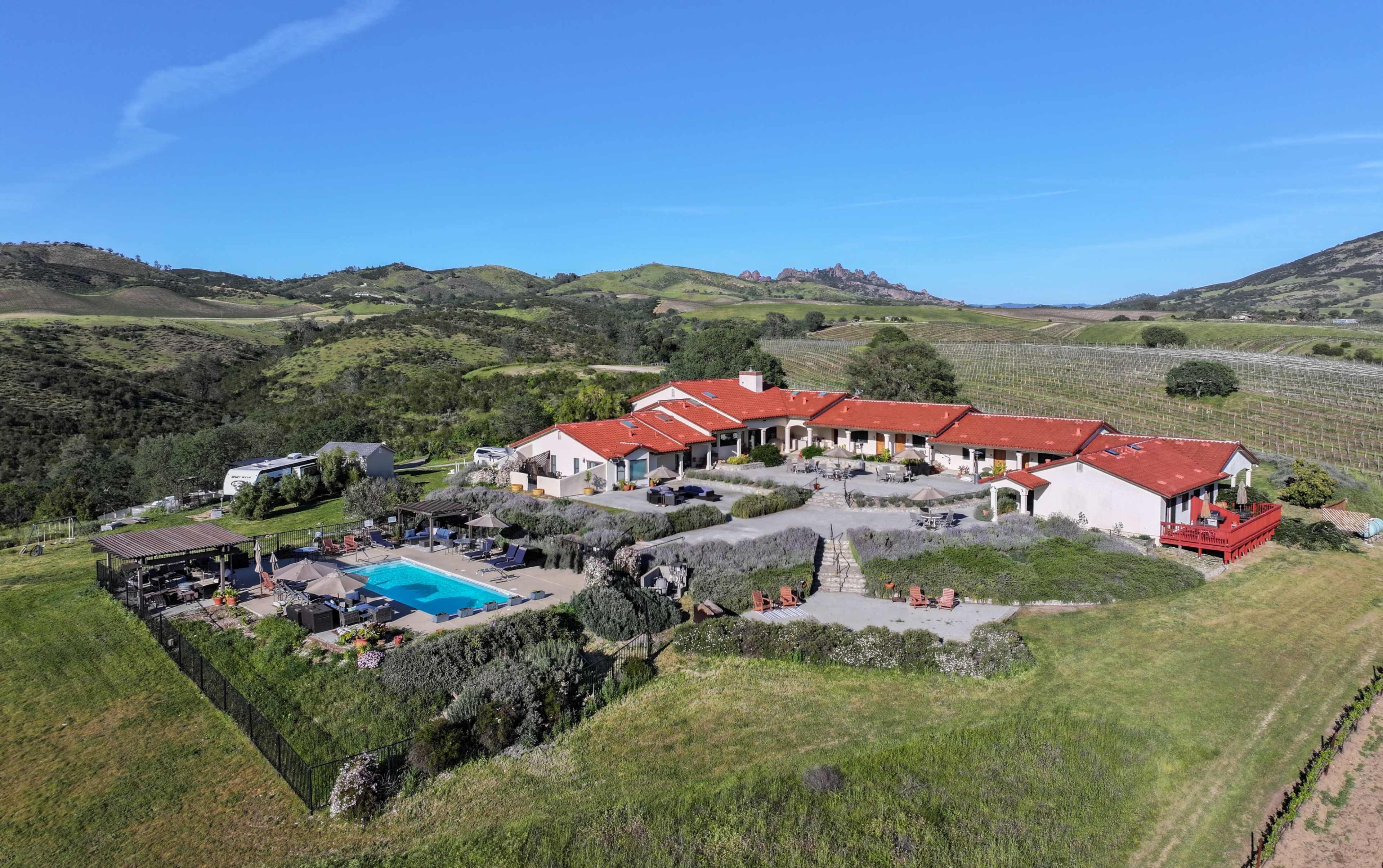 The image shows a large villa with a red-tiled roof set against a backdrop of rolling green hills and vineyards.