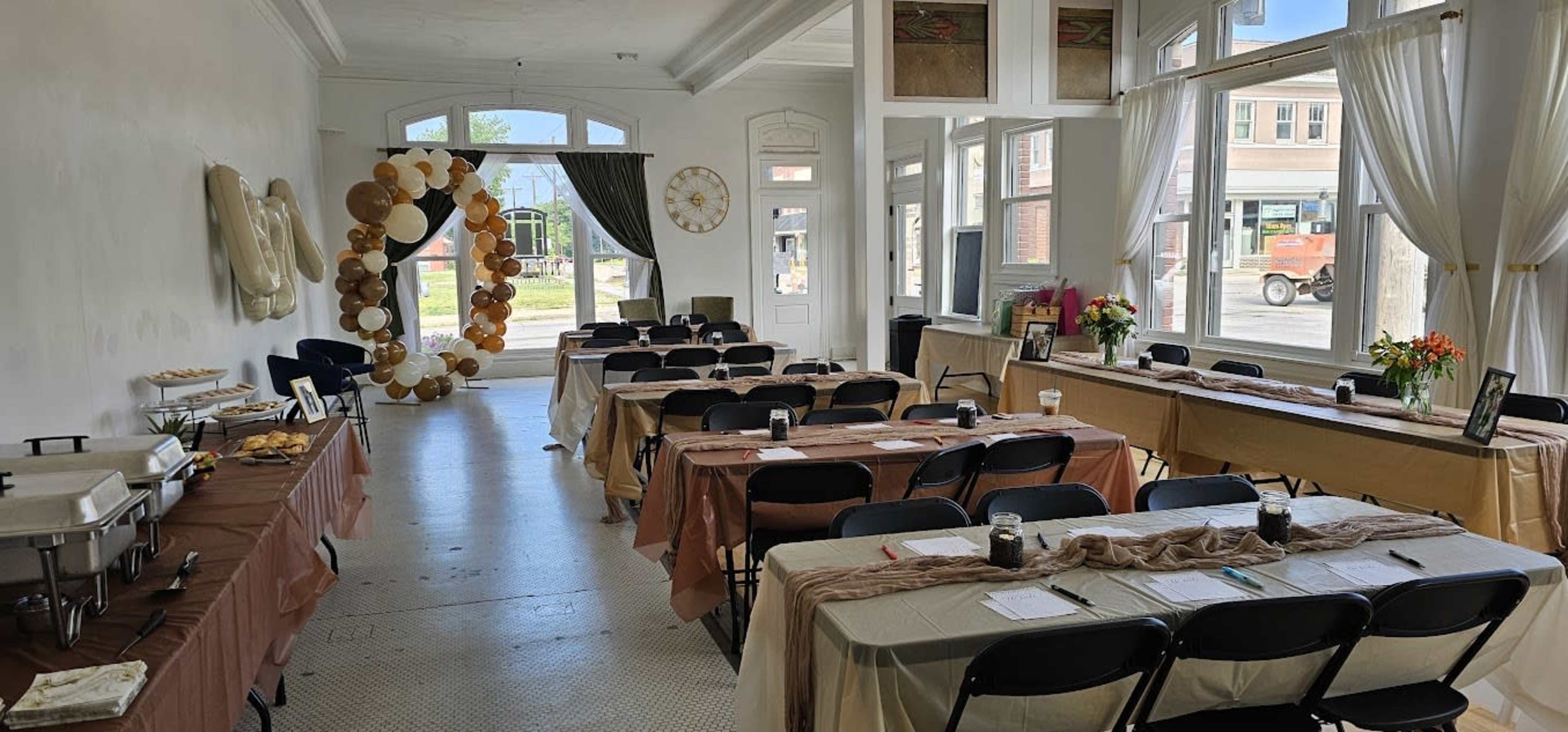 The image shows a large room set up for an event, featuring multiple tables with chairs arranged in rows, adorned with tablecloths and decorative elements, including a balloon arch near the entrance.