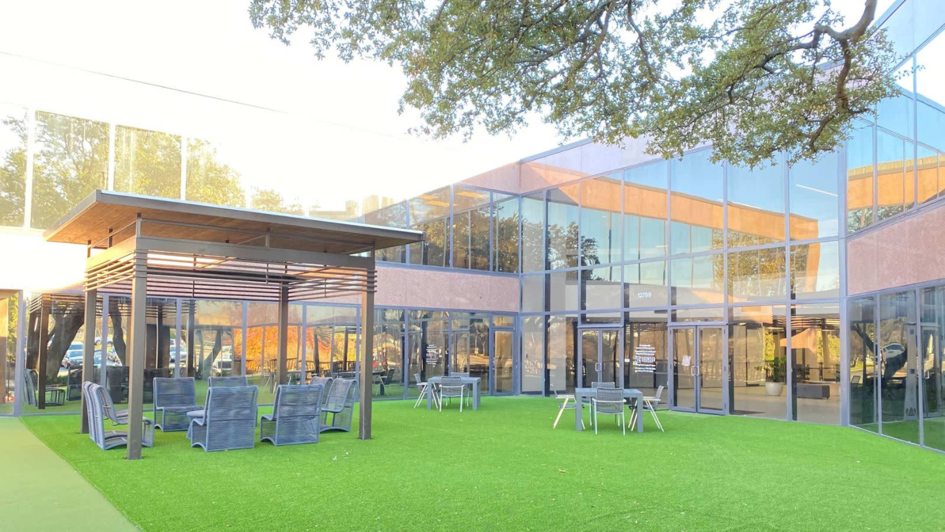 A modern outdoor seating area with gray furniture set on green artificial grass, surrounded by large glass buildings.