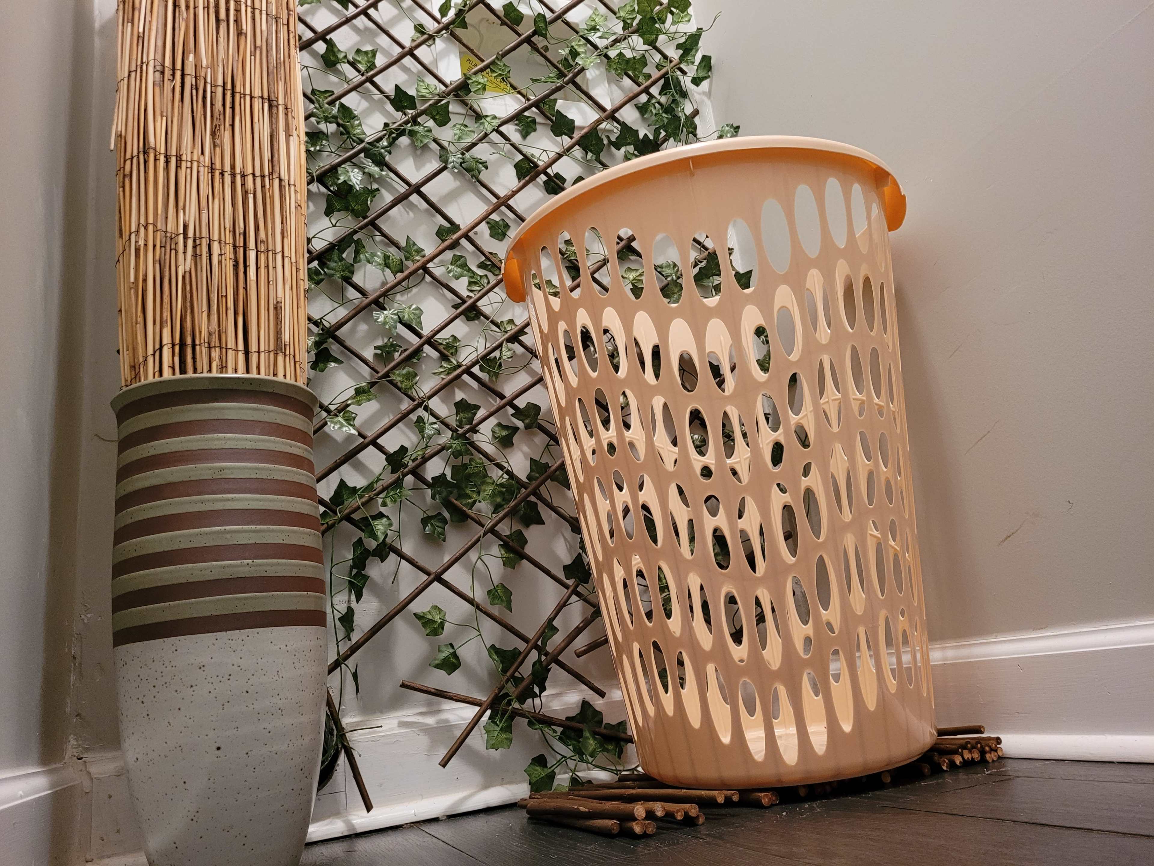 A plastic laundry basket rests beside a tall decorative vase and a trellis covered in green foliage against a wall.