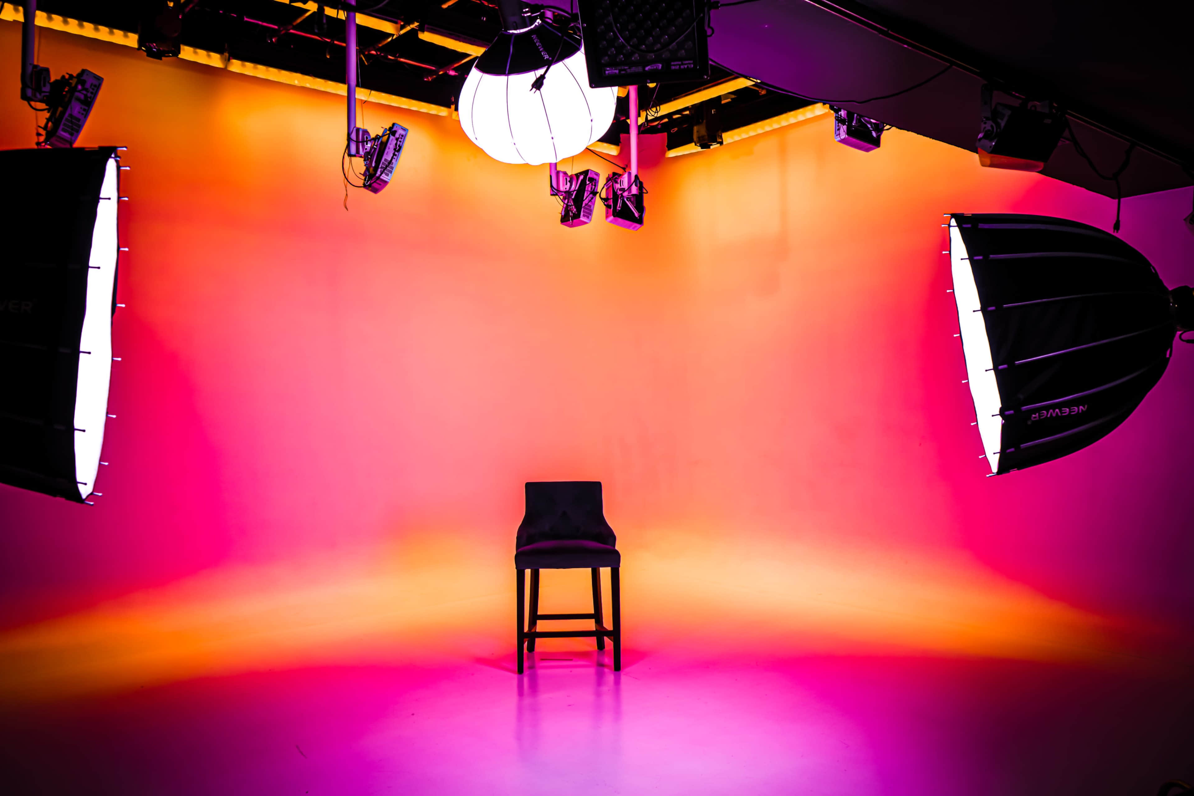 A single black chair is centered in a brightly lit studio with an orange and pink gradient backdrop.