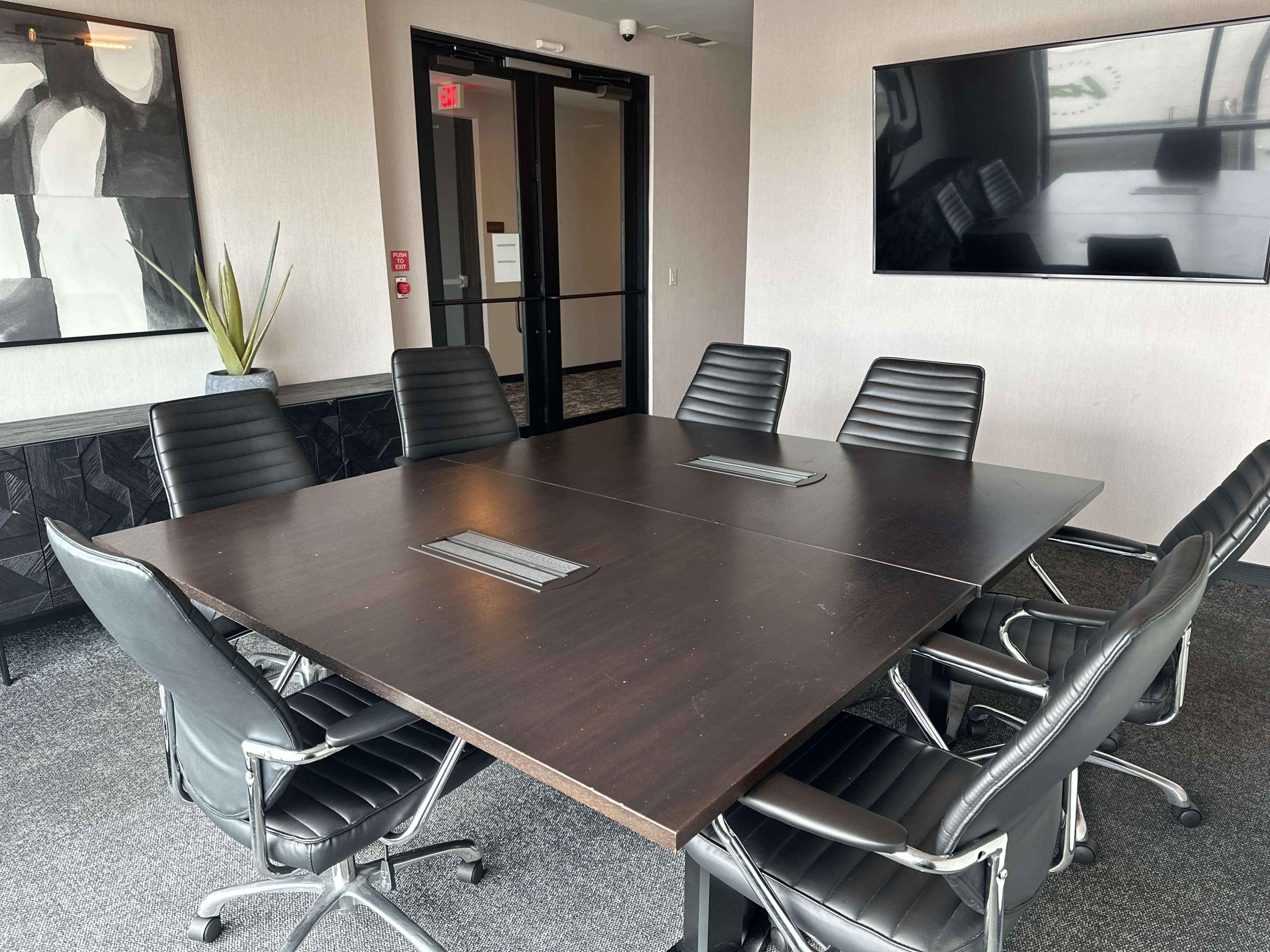 A modern conference room features a large dark wooden table surrounded by eight black leather chairs, with a large screen mounted on the wall and a plant in the corner.