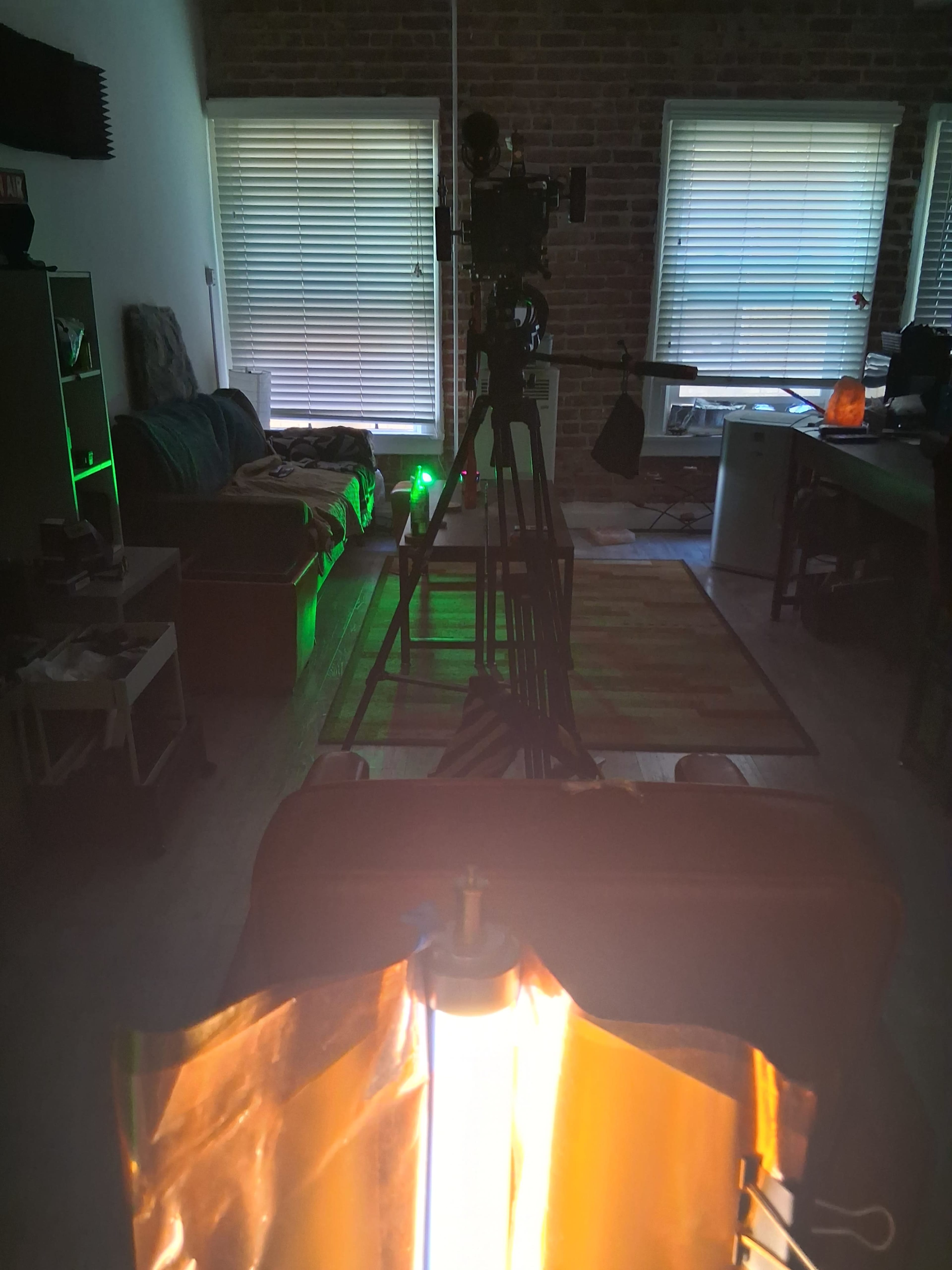 A video camera is set up on a tripod in a dimly lit room with green LED lighting and visible furniture, creating a filming environment.