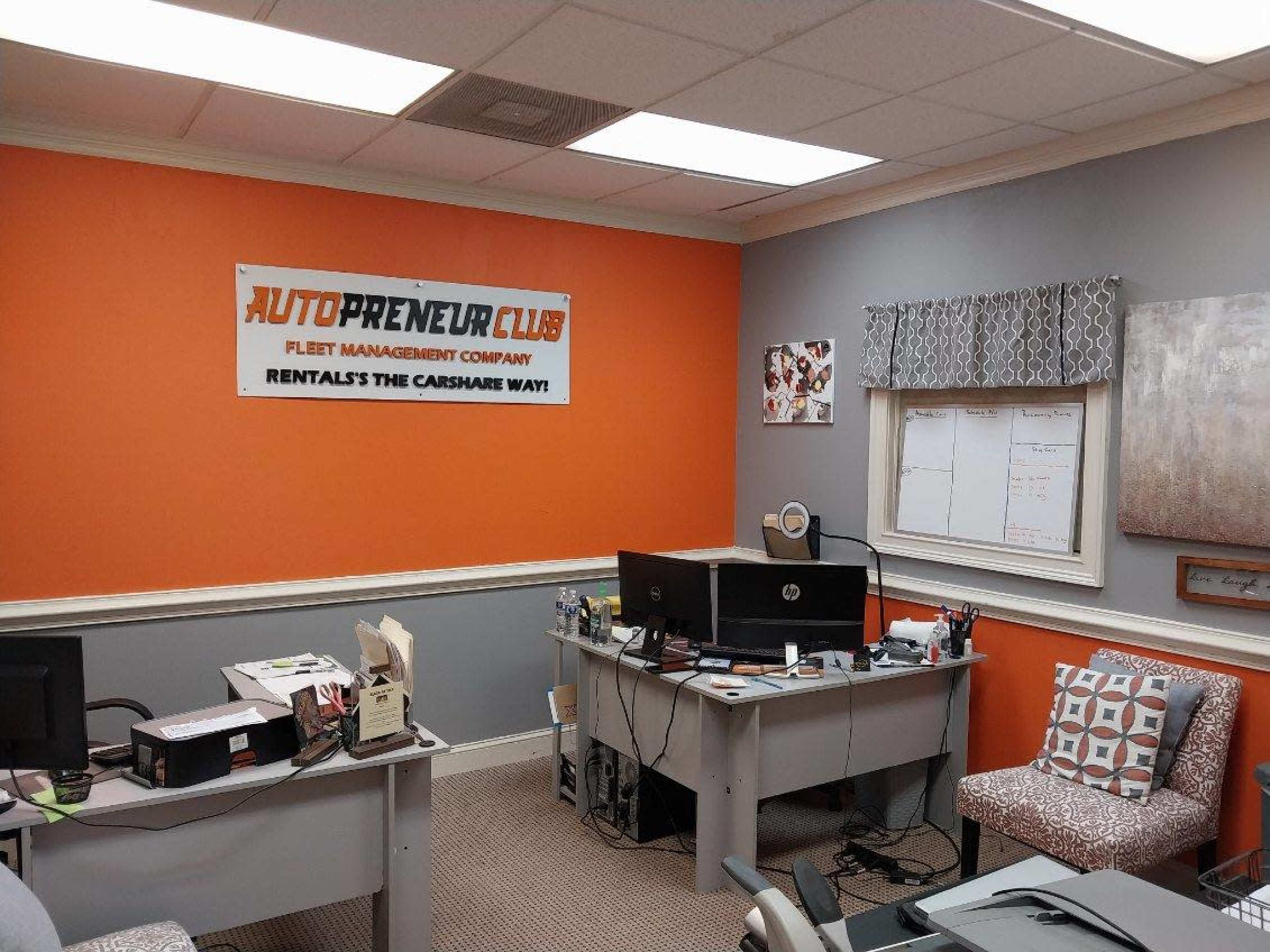 An office space with orange and gray walls, featuring a desk with a computer and office supplies, along with a large banner for "AutoPreneur Club" on the wall.